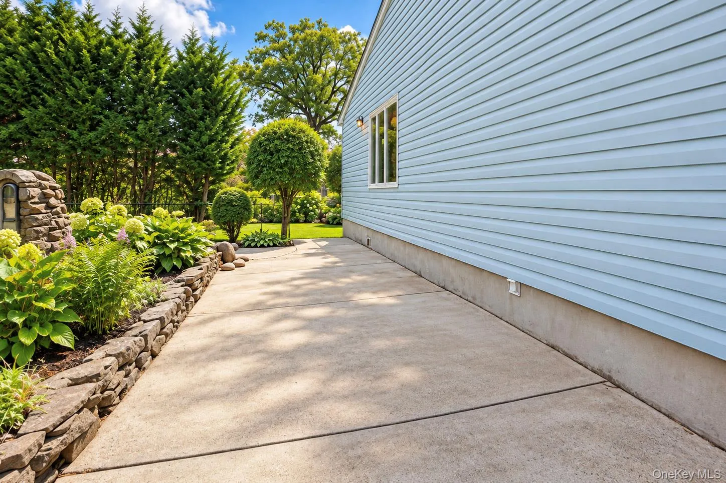 Exterior featuring light blue siding, a window with white trim, a concrete walkway, a stone-retained garden bed with varied plantings, and mature landscaping Exterior featuring light blue siding, a window with white trim, a concrete walkway, a stone-retained garden bed with varied plantings, and mature landscaping