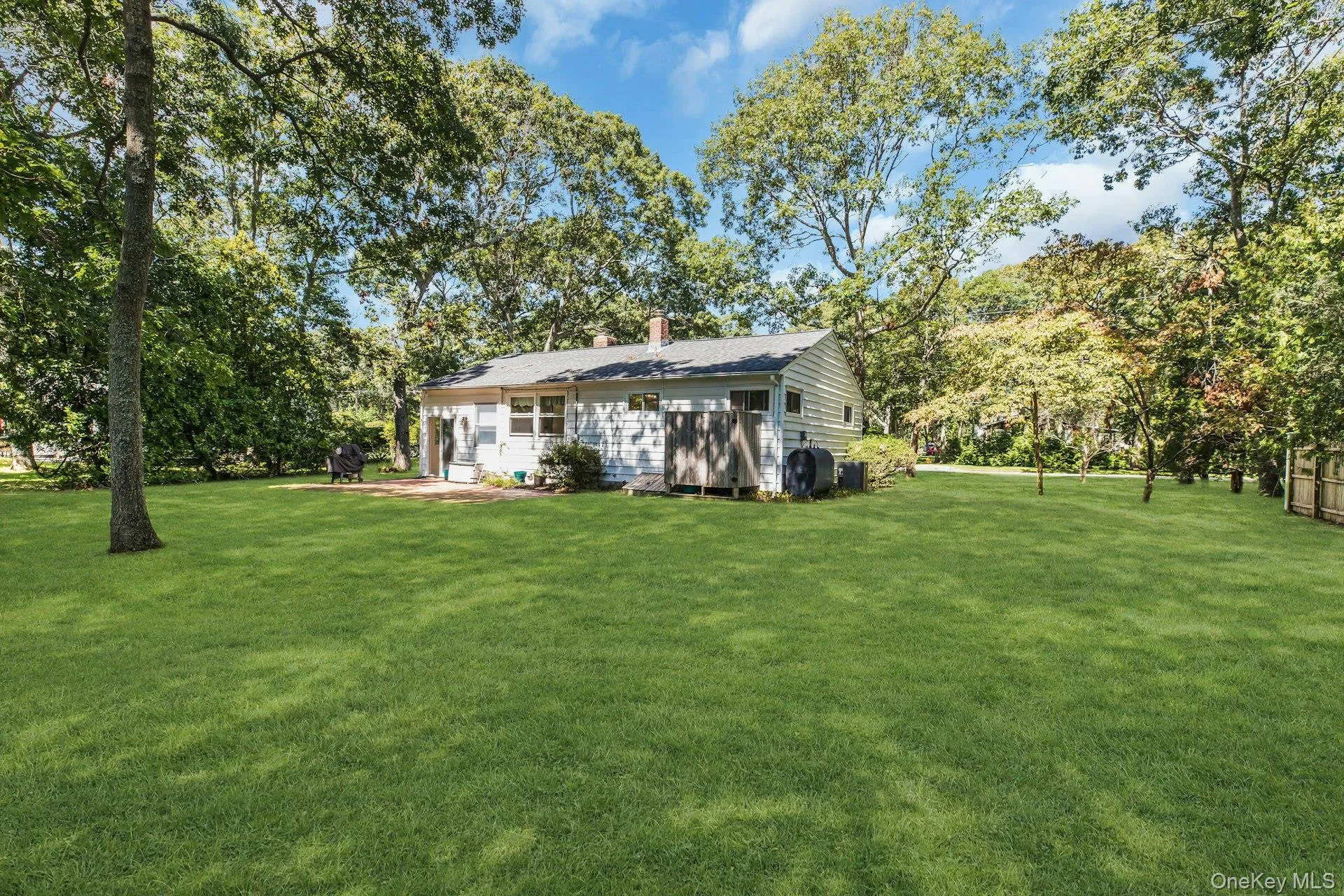 Expansive green lawn, mature trees, white siding exterior, shingle roof, and a paved patio area Expansive green lawn, mature trees, white siding exterior, shingle roof, and a paved patio area