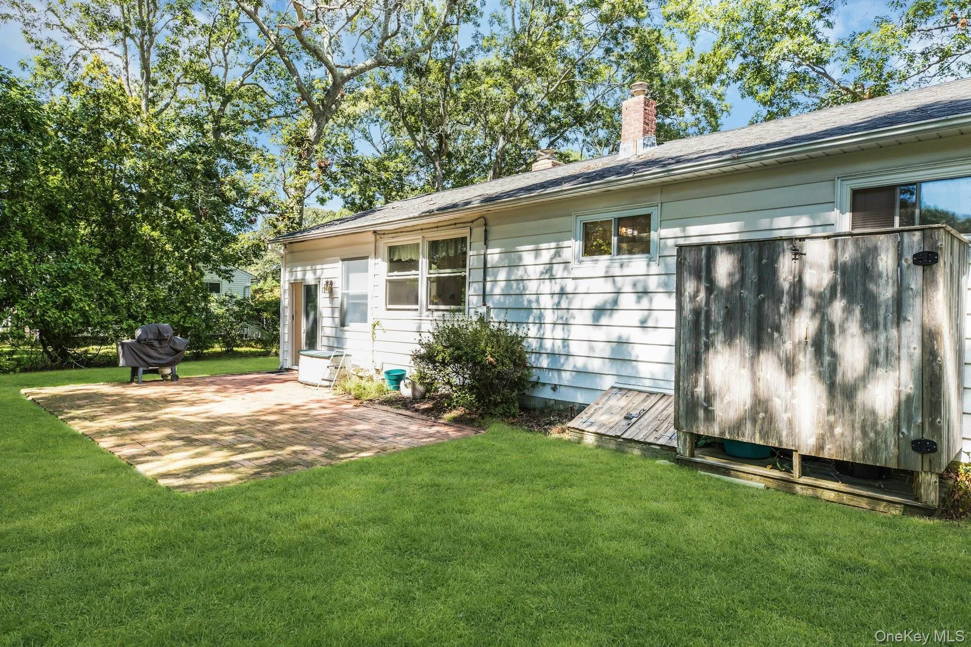 Expansive green lawn, brick paver patio, mature trees, white horizontal siding, and a red brick chimney Expansive green lawn, brick paver patio, mature trees, white horizontal siding, and a red brick chimney