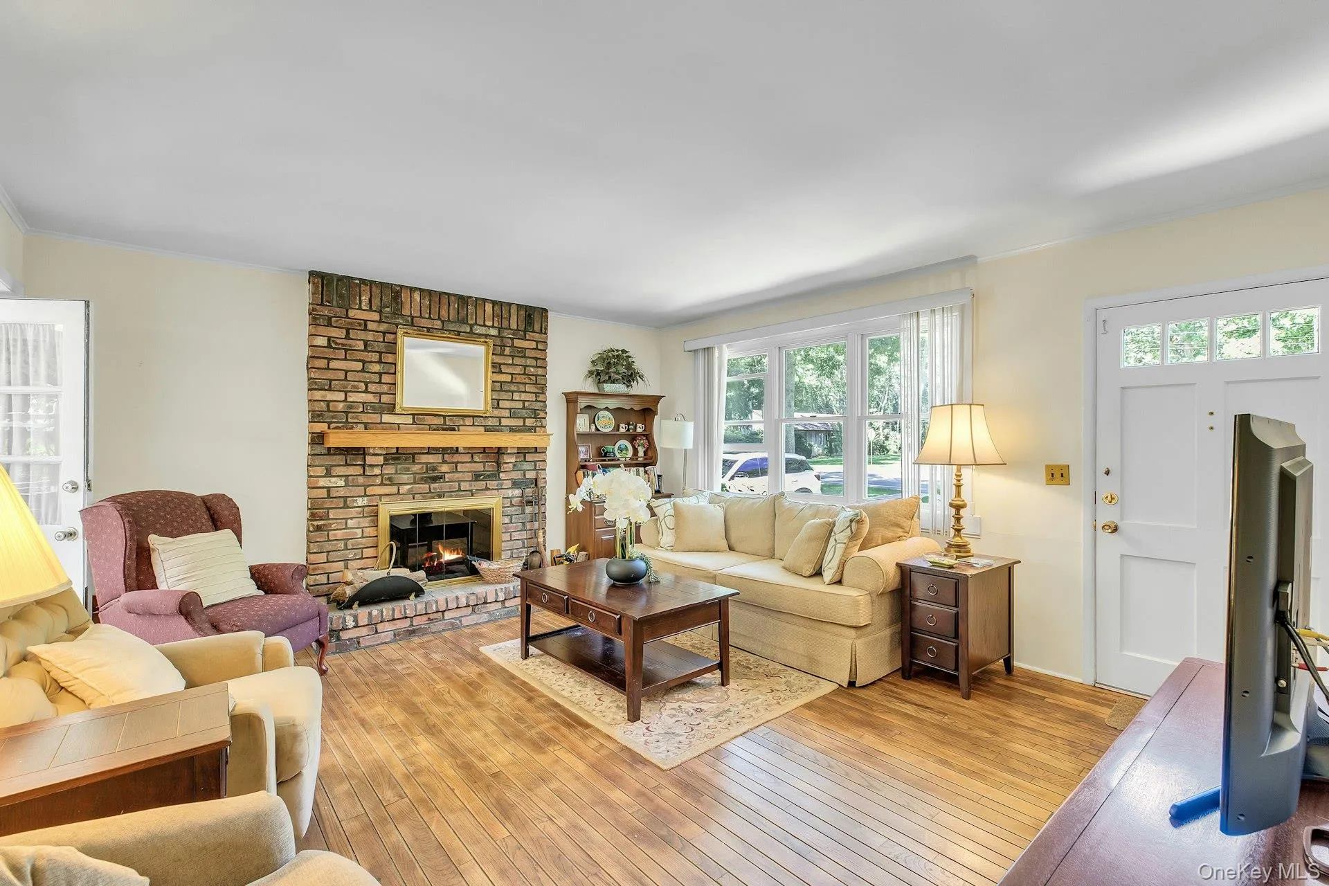 Living room featuring wood-finish flooring, a brick fireplace with a wood mantle, a multi-pane window, and a white entry door with upper glass panels Living room featuring wood-finish flooring, a brick fireplace with a wood mantle, a multi-pane window, and a white entry door with upper glass panels