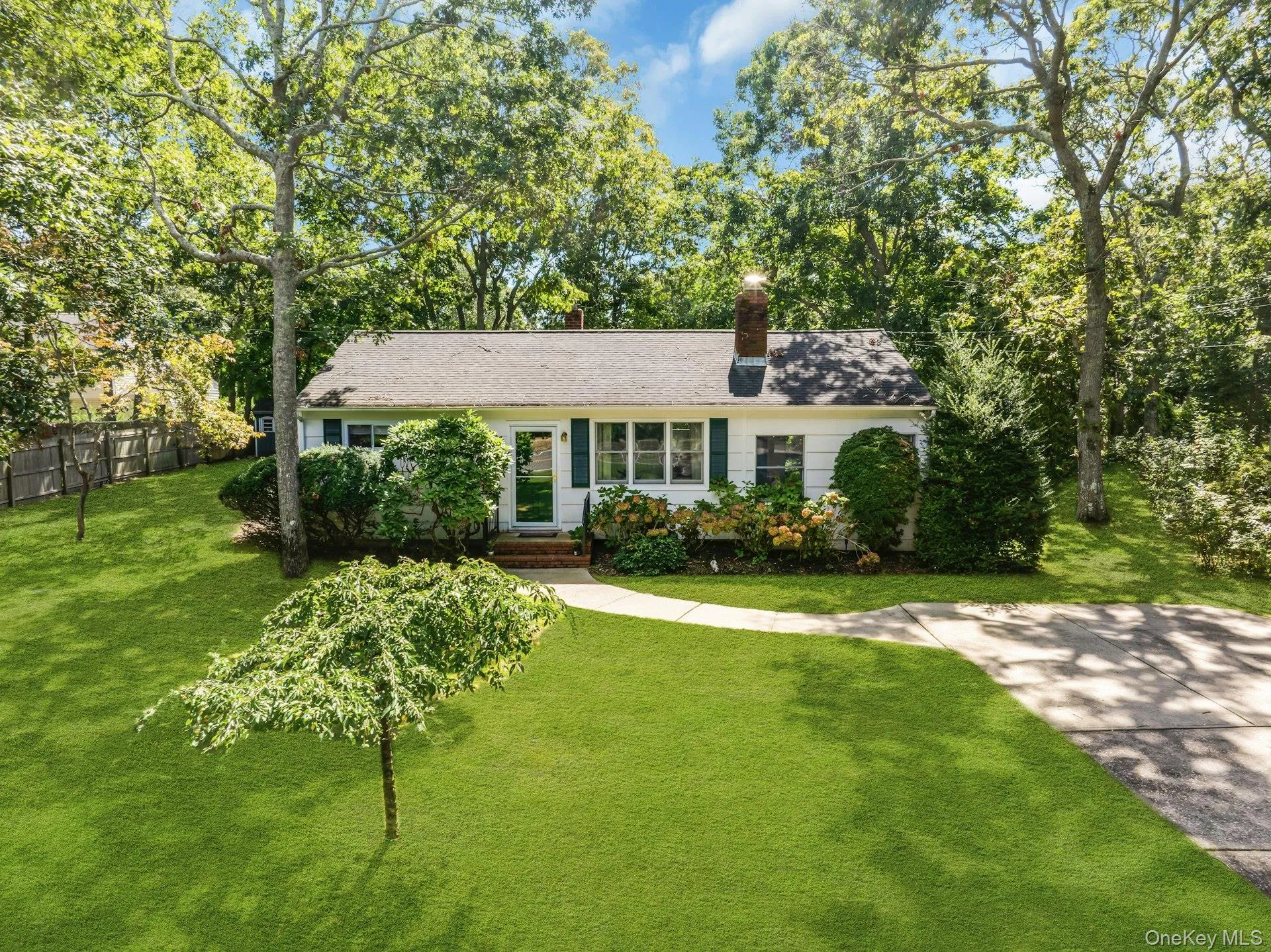 White exterior with dark green shutters, a paved driveway, a concrete pathway, a shingle roof, and mature trees White exterior with dark green shutters, a paved driveway, a concrete pathway, a shingle roof, and mature trees