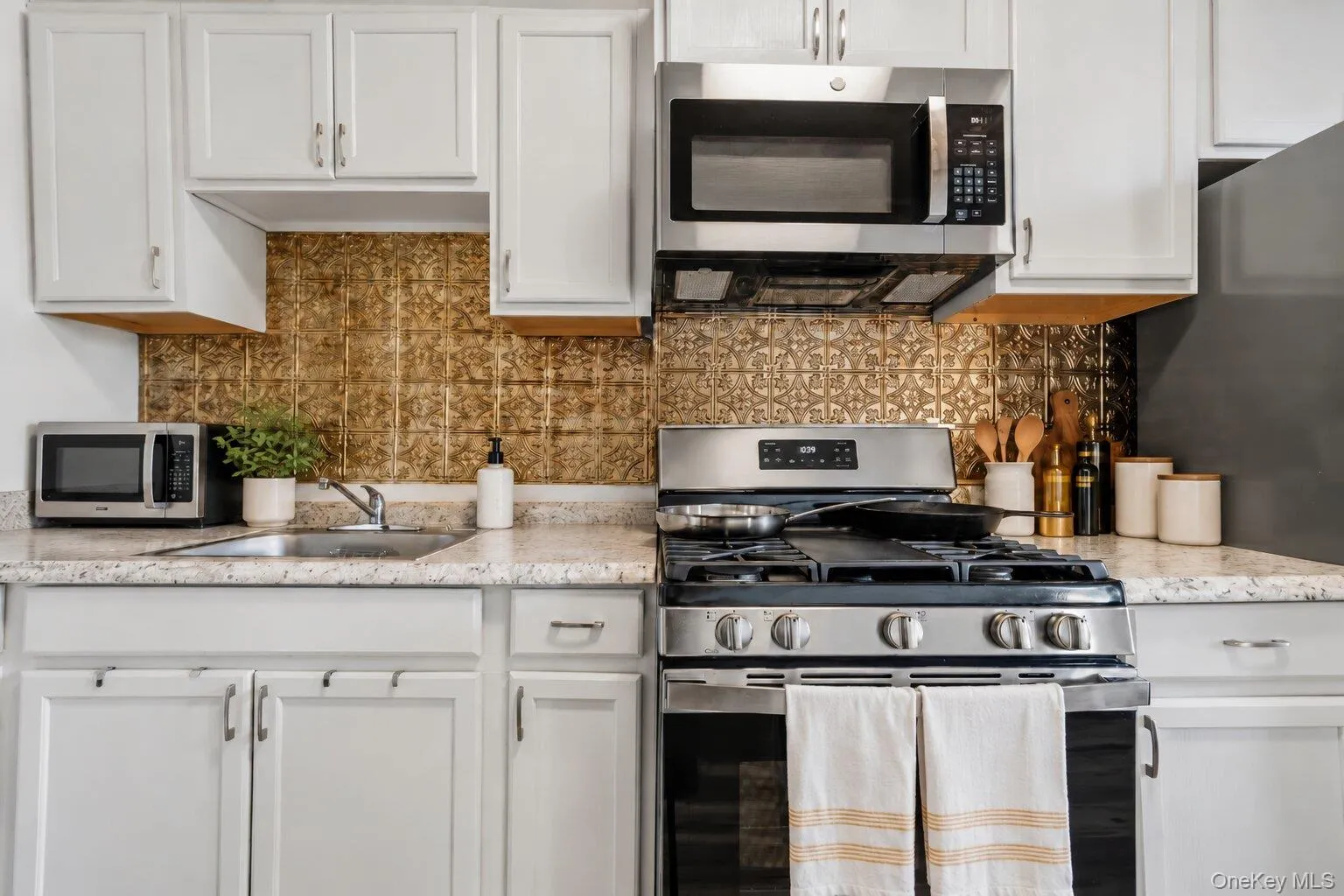 Virtually Staged Furniture. Close view of the kitchen showcasing white cabinetry, stainless steel gas range and microwave, double-basin sink, decorative metallic backsplash, and countertop workspace. Virtually Staged Furniture. Close view of the kitchen showcasing white cabinetry, stainless steel gas range and microwave, double-basin sink, decorative metallic backsplash, and countertop workspace.