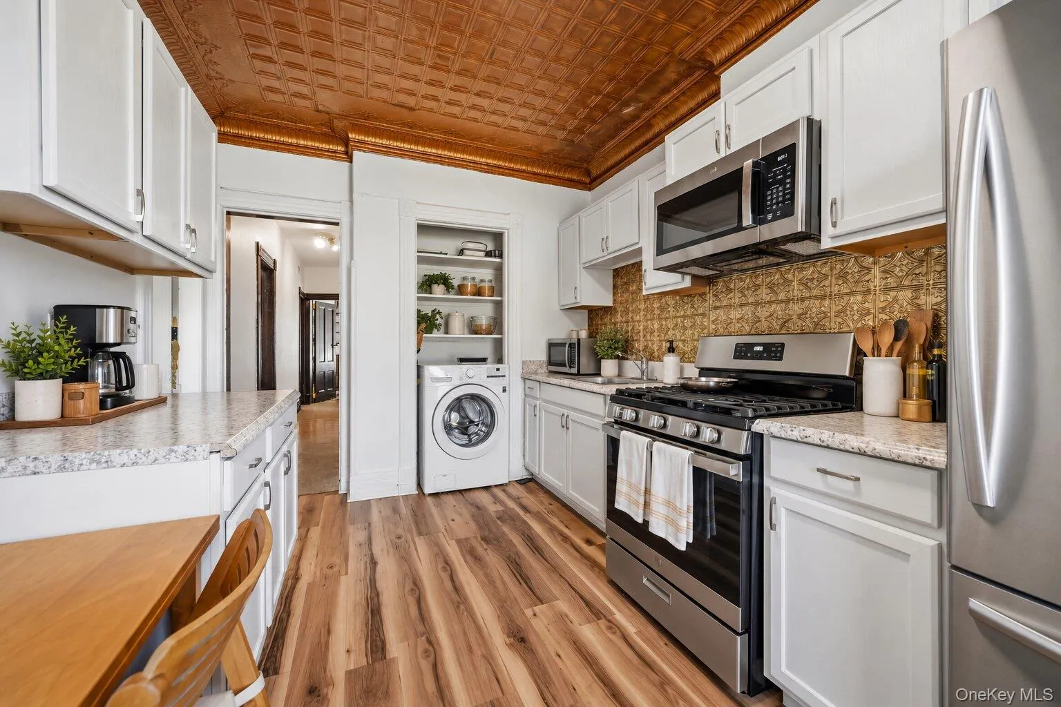 Virtually Staged Furniture. Spacious kitchen with white cabinetry, stainless steel appliances, gas range, decorative backsplash, wood-look flooring, laundry area, and copper-toned patterned ceiling. Virtually Staged Furniture. Spacious kitchen with white cabinetry, stainless steel appliances, gas range, decorative backsplash, wood-look flooring, laundry area, and copper-toned patterned ceiling.