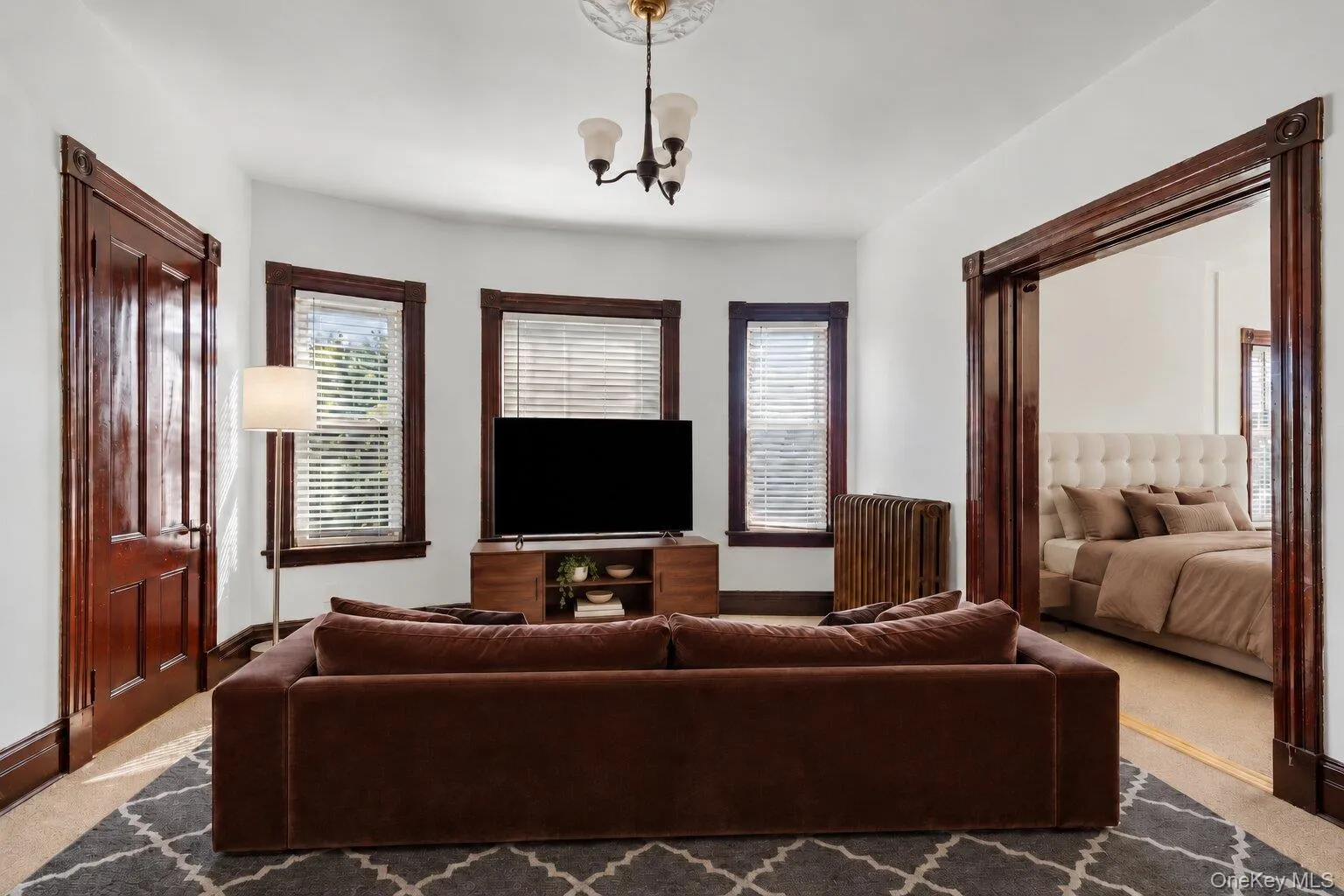 Virtually Staged Furniture. Living room with multiple windows, radiator, dark wood trim, and an adjoining bedroom visible through the doorway. Virtually Staged Furniture. Living room with multiple windows, radiator, dark wood trim, and an adjoining bedroom visible through the doorway.