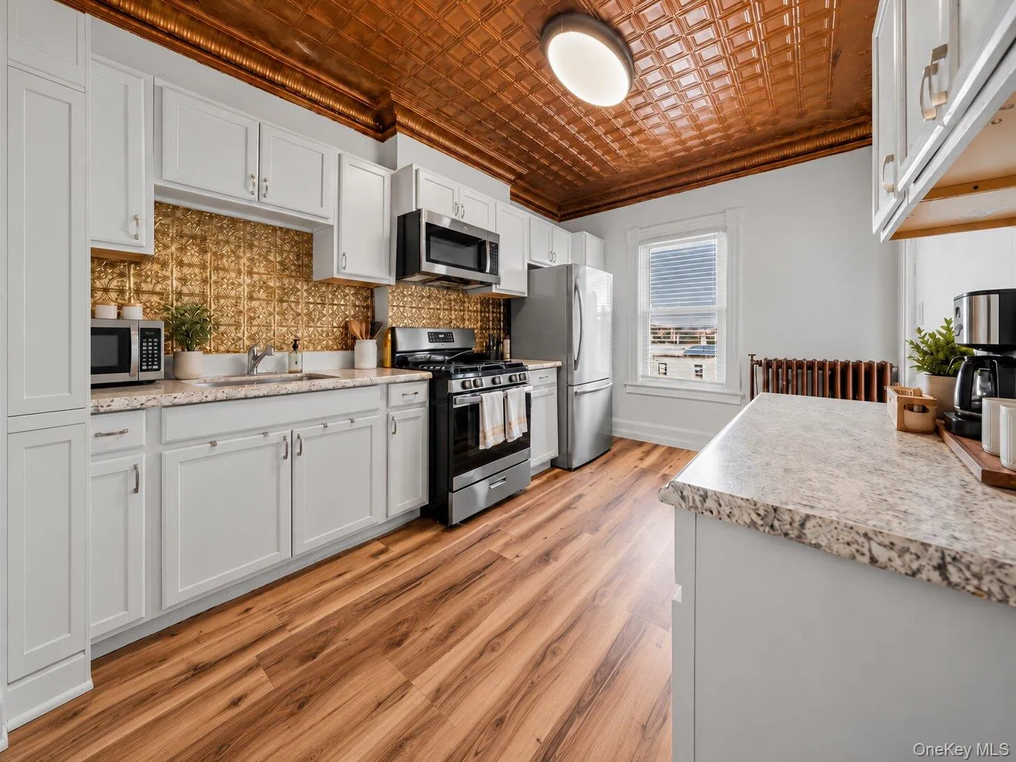 Virtually Staged Furniture. Bright kitchen featuring white cabinetry, granite-look countertops, stainless steel appliances, wood-look flooring, decorative metallic backsplash, and a distinctive copper-toned patterned ceiling. Virtually Staged Furniture. Bright kitchen featuring white cabinetry, granite-look countertops, stainless steel appliances, wood-look flooring, decorative metallic backsplash, and a distinctive copper-toned patterned ceiling.