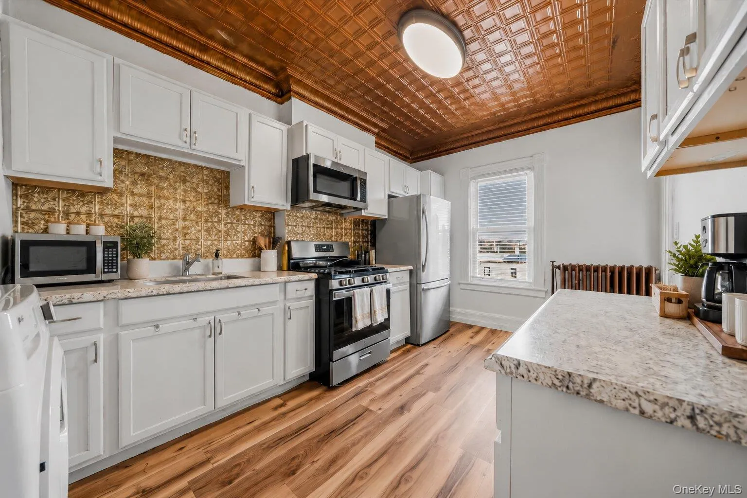 Virtually Staged Furniture. Bright kitchen featuring white cabinetry, granite-look countertops, stainless steel appliances, wood-look flooring, decorative metallic backsplash, and a distinctive copper-toned patterned ceiling. Virtually Staged Furniture. Bright kitchen featuring white cabinetry, granite-look countertops, stainless steel appliances, wood-look flooring, decorative metallic backsplash, and a distinctive copper-toned patterned ceiling.