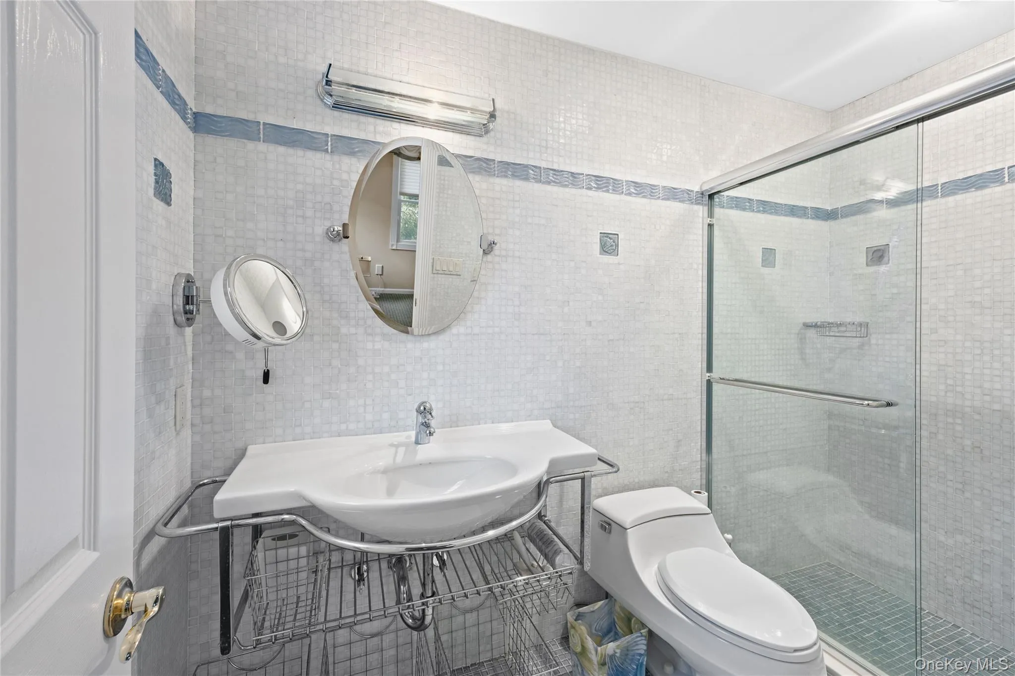 Shower with glass enclosure, white square tile walls with blue accent banding, white pedestal sink with chrome-finish basin, and oval wall mirror Shower with glass enclosure, white square tile walls with blue accent banding, white pedestal sink with chrome-finish basin, and oval wall mirror