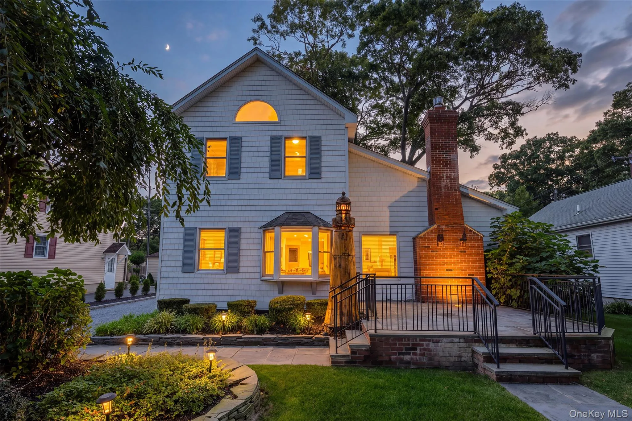 White shingle siding with gray shutters, bay window, prominent brick chimney, multi-level patio with black railings, and stone-edged landscaping with path lighting White shingle siding with gray shutters, bay window, prominent brick chimney, multi-level patio with black railings, and stone-edged landscaping with path lighting