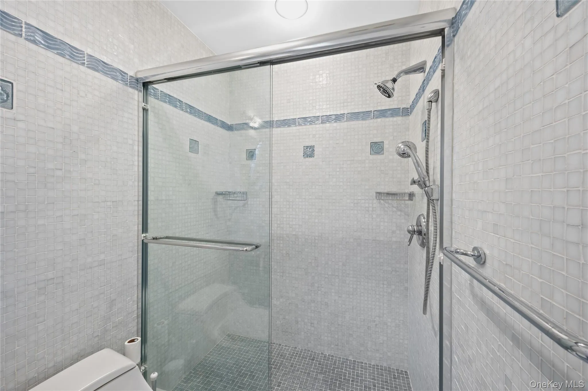 Glass-enclosed shower with a handheld shower fixture, overhead showerhead, and mosaic tile walls Glass-enclosed shower with a handheld shower fixture, overhead showerhead, and mosaic tile walls