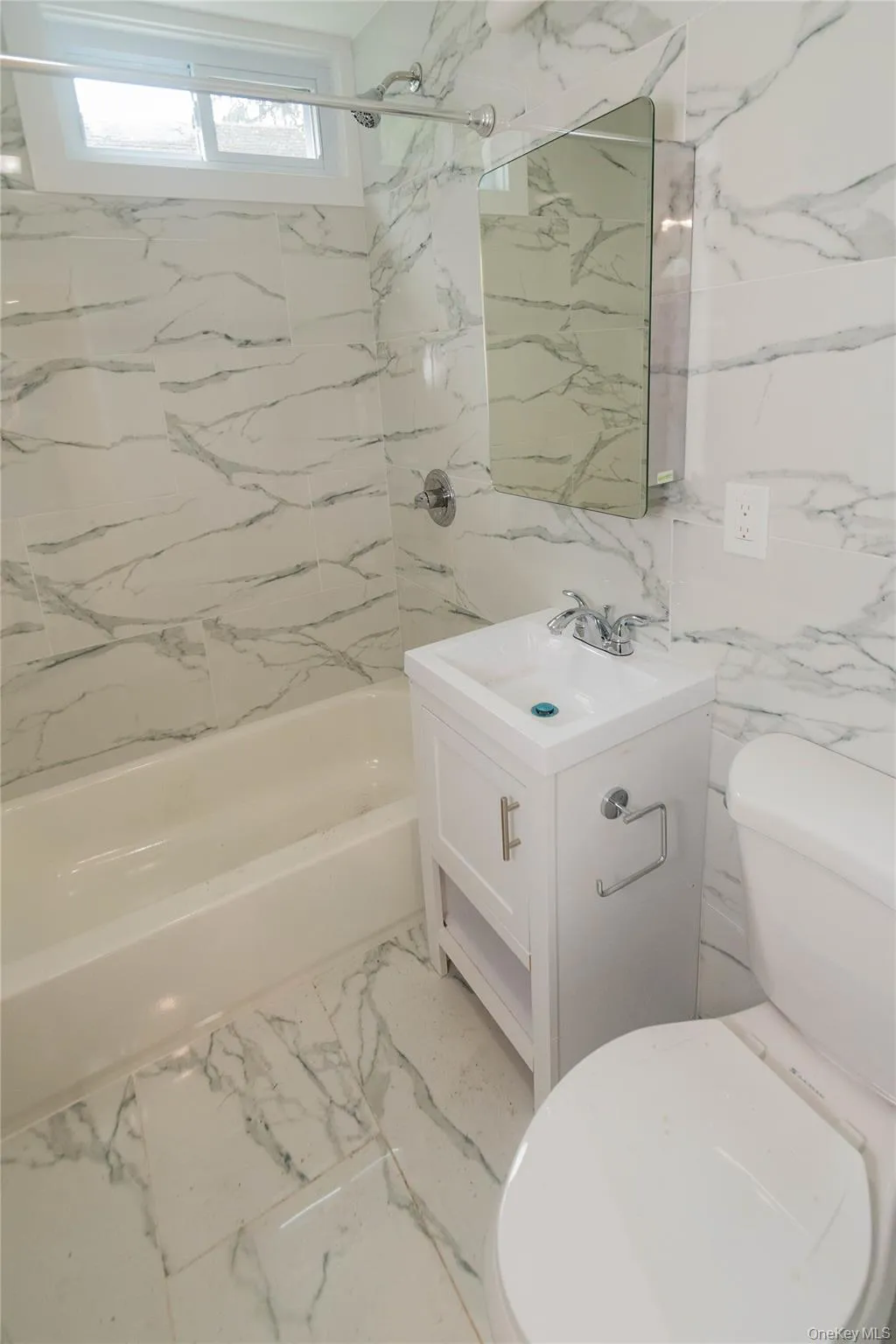 Full bathroom featuring a white vanity with integrated sink, chrome fixtures, rectangular wall mirror, and floor-to-ceiling marbled tile Full bathroom featuring a white vanity with integrated sink, chrome fixtures, rectangular wall mirror, and floor-to-ceiling marbled tile