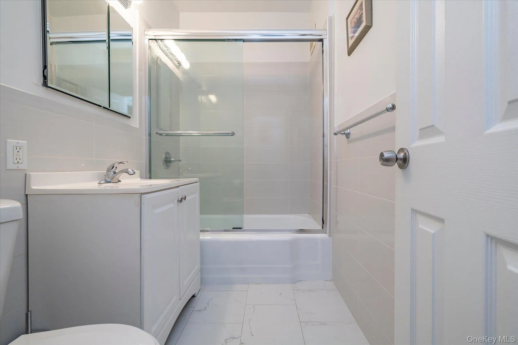 Bathroom featuring a single vanity with storage, a shower-tub combination with glass enclosure, white wall tiling, and white floor tiling Bathroom featuring a single vanity with storage, a shower-tub combination with glass enclosure, white wall tiling, and white floor tiling