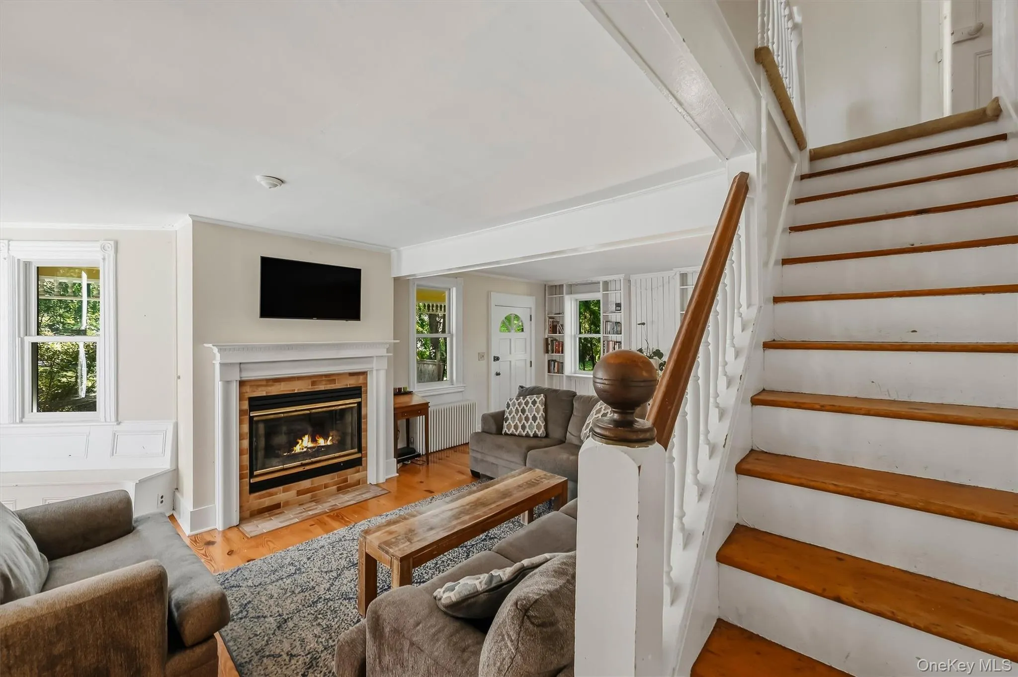 Hardwood flooring, brick fireplace with a white mantel, white trim, recessed lighting, and a detailed staircase with a wood handrail Hardwood flooring, brick fireplace with a white mantel, white trim, recessed lighting, and a detailed staircase with a wood handrail