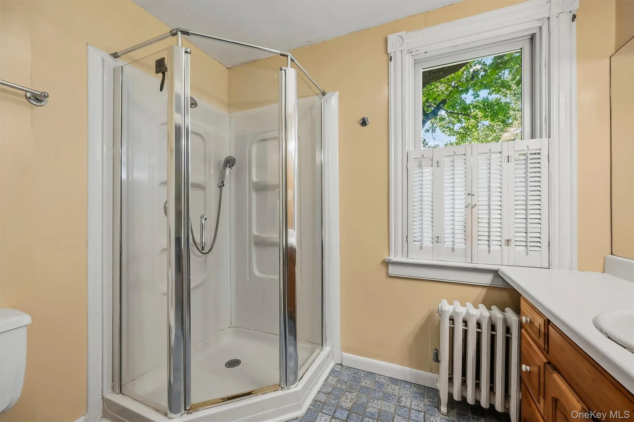 Corner shower enclosure with glass panels, white window shutters, and a wood-finish vanity with an integrated sink Corner shower enclosure with glass panels, white window shutters, and a wood-finish vanity with an integrated sink