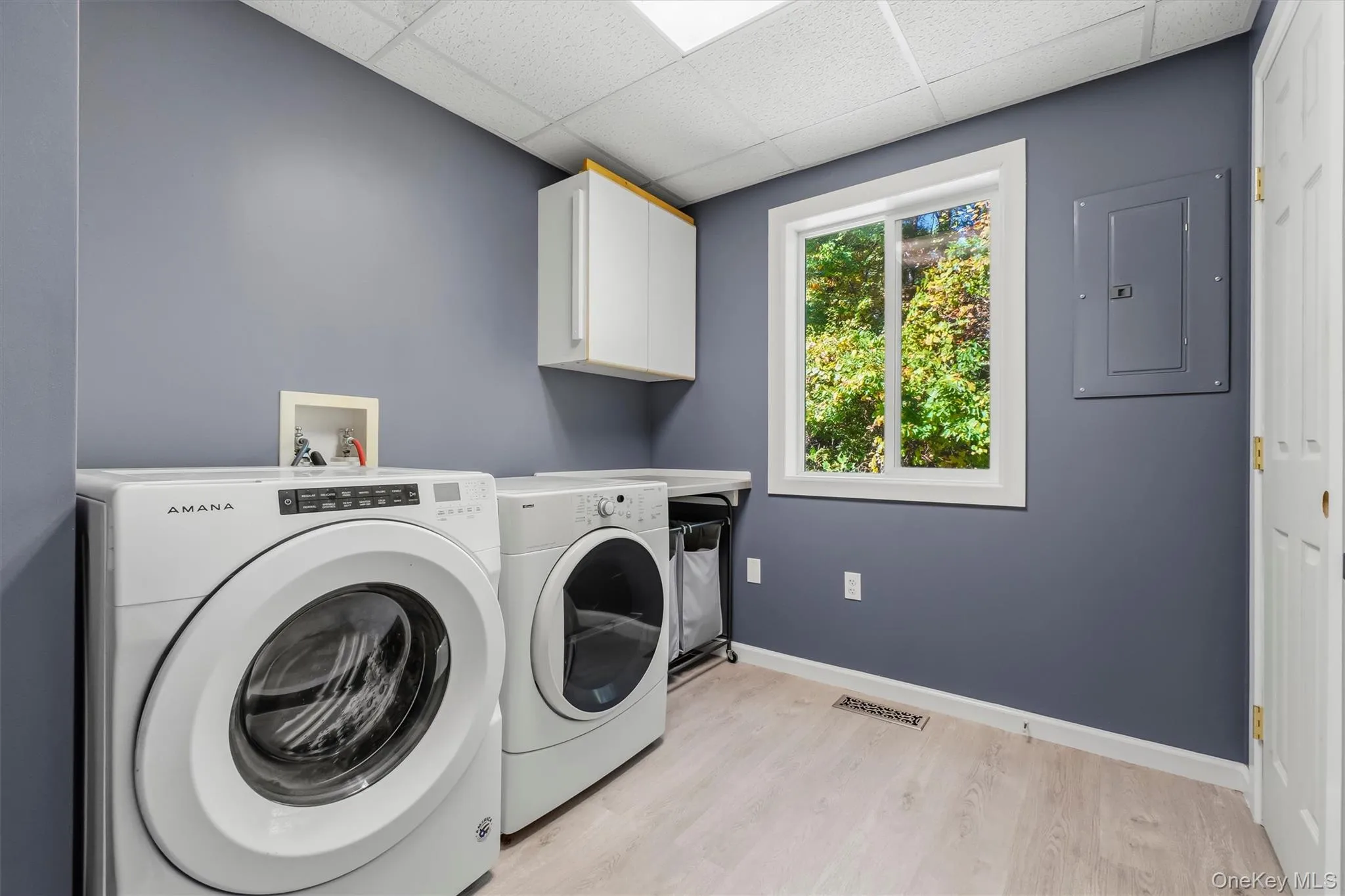 Spacious laundry room featuring wood-finish flooring, white cabinetry, a large window, and a built-in folding counter Spacious laundry room featuring wood-finish flooring, white cabinetry, a large window, and a built-in folding counter
