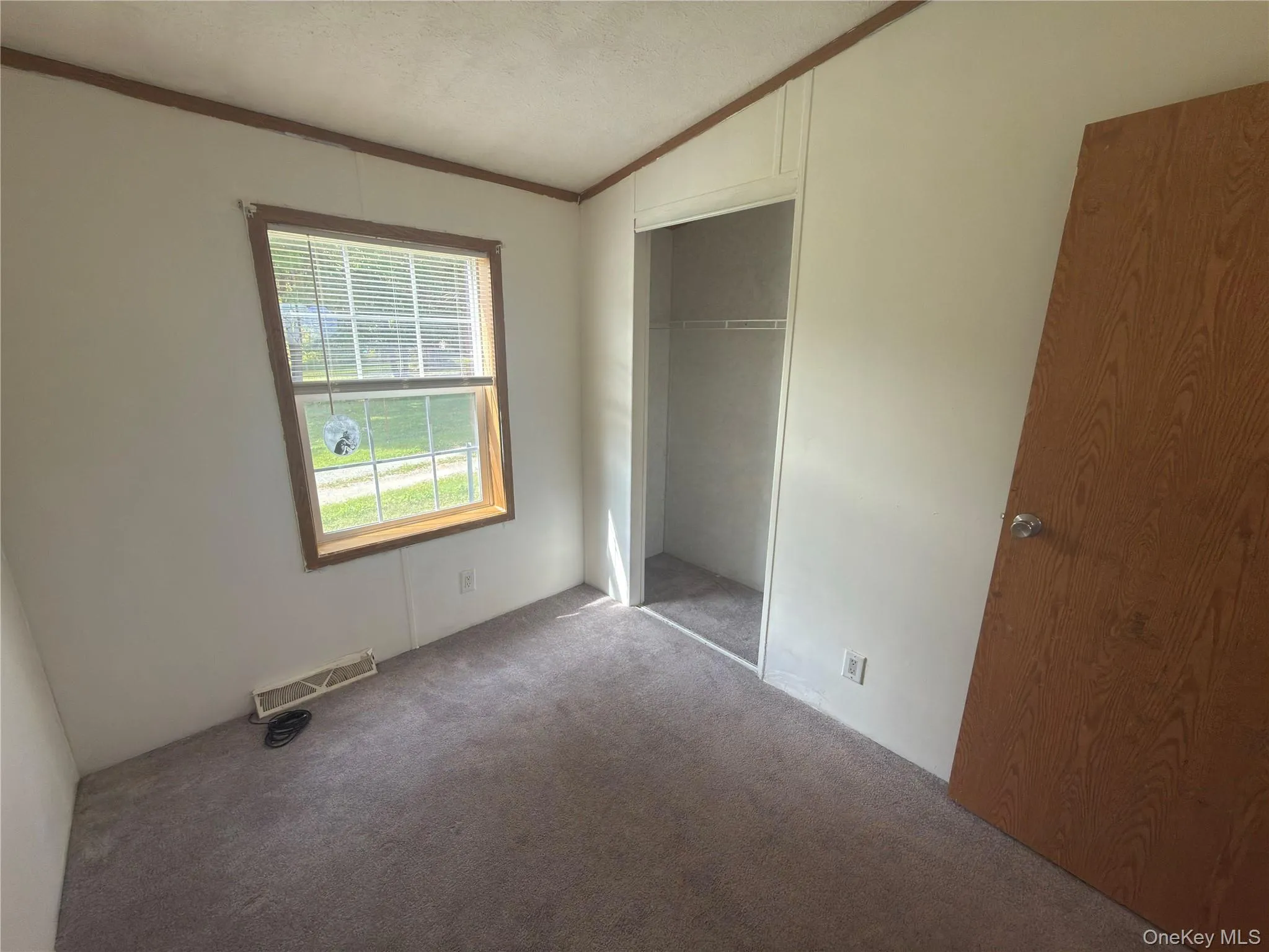 Recessed closet with a built-in shelf, light-colored carpeting, a window with horizontal blinds, and a wood-finish door Recessed closet with a built-in shelf, light-colored carpeting, a window with horizontal blinds, and a wood-finish door