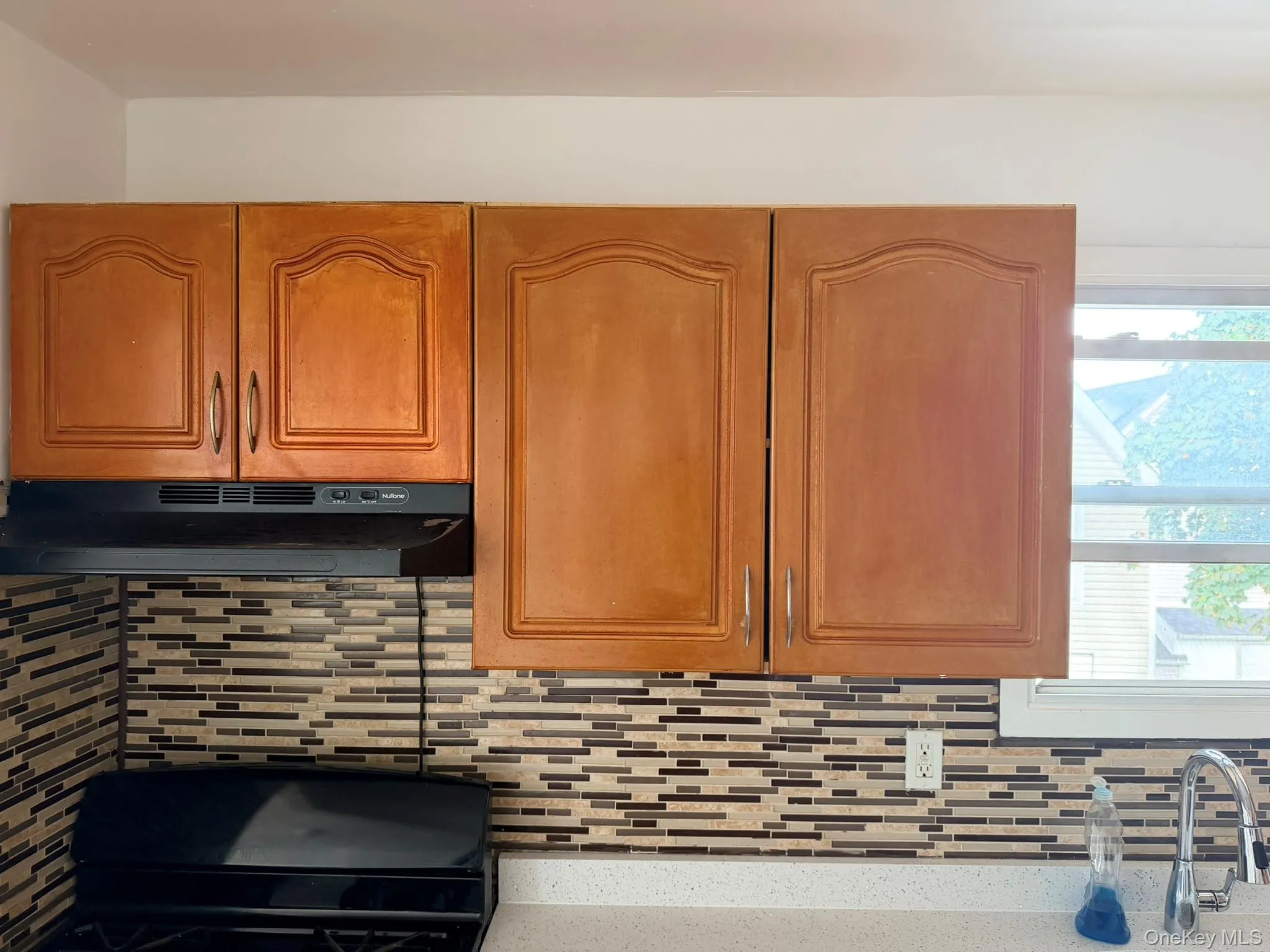 Recessed panel cabinetry with arched details, mosaic tile backsplash, built-in range hood, solid surface countertop, and chrome gooseneck faucet Recessed panel cabinetry with arched details, mosaic tile backsplash, built-in range hood, solid surface countertop, and chrome gooseneck faucet