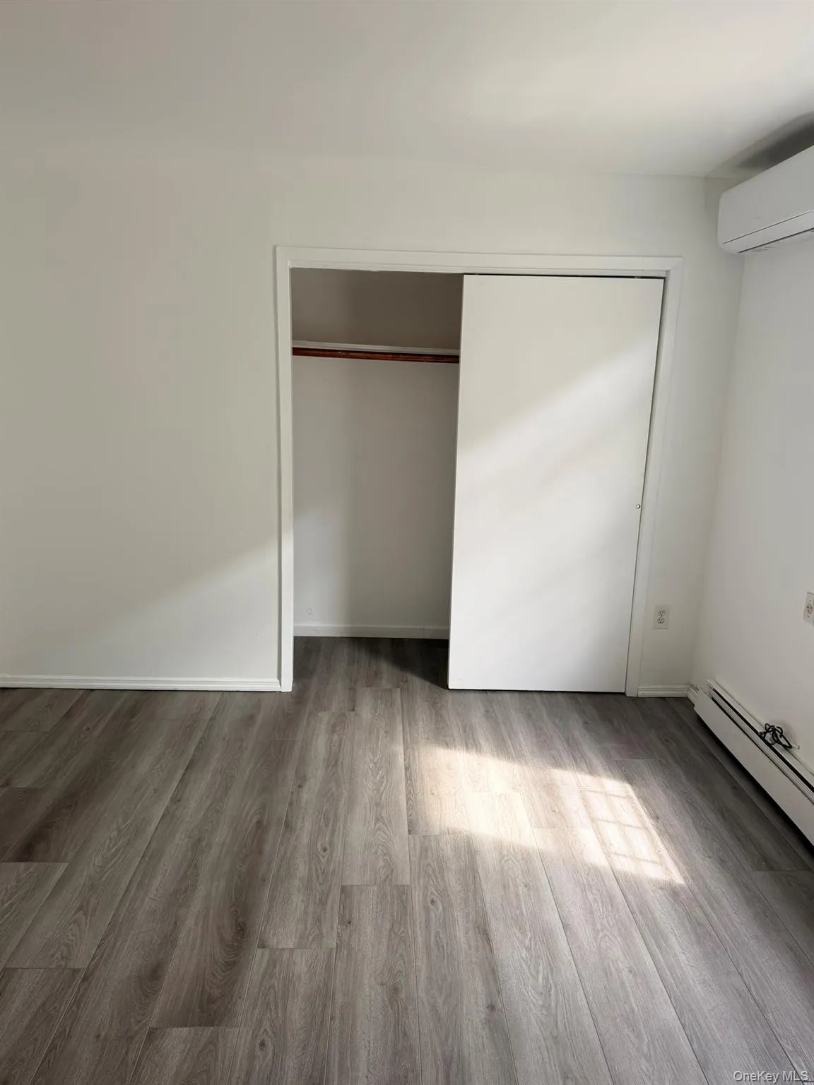 Wood-finish flooring and white walls, featuring a closet with sliding doors and a wall-mounted air conditioning unit Wood-finish flooring and white walls, featuring a closet with sliding doors and a wall-mounted air conditioning unit
