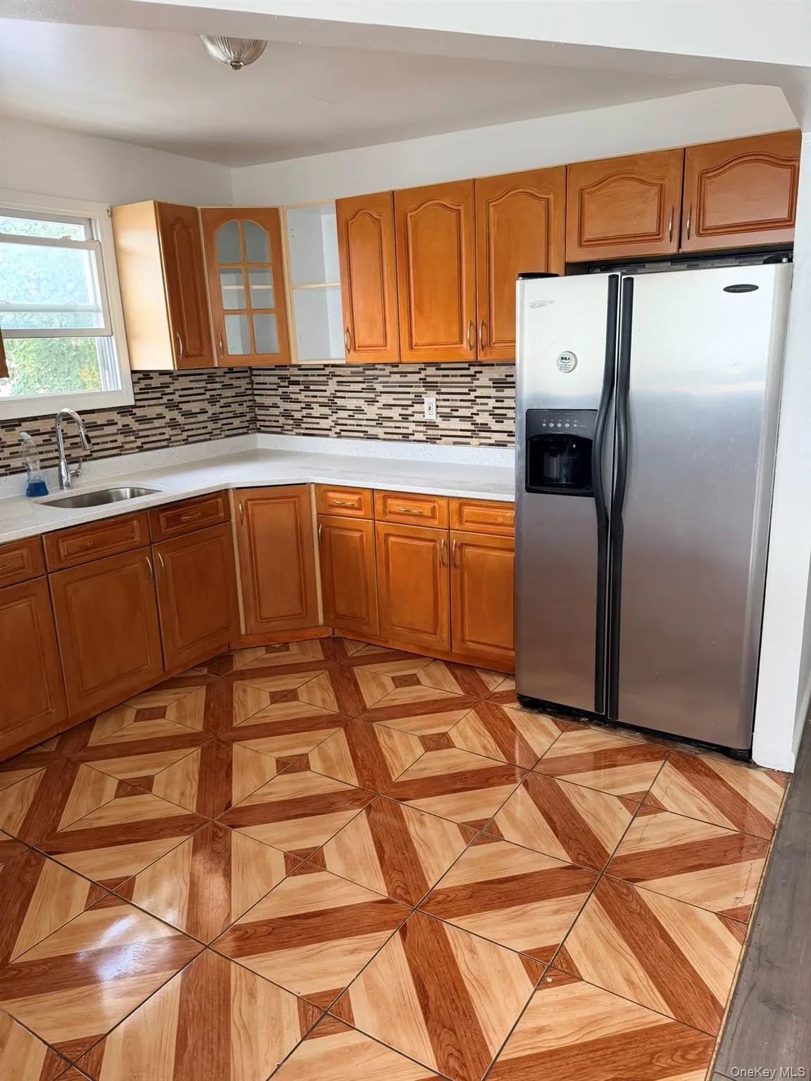 Wood-tone cabinetry, stainless steel refrigerator, white countertops, mosaic tile backsplash, and patterned floor tiling Wood-tone cabinetry, stainless steel refrigerator, white countertops, mosaic tile backsplash, and patterned floor tiling