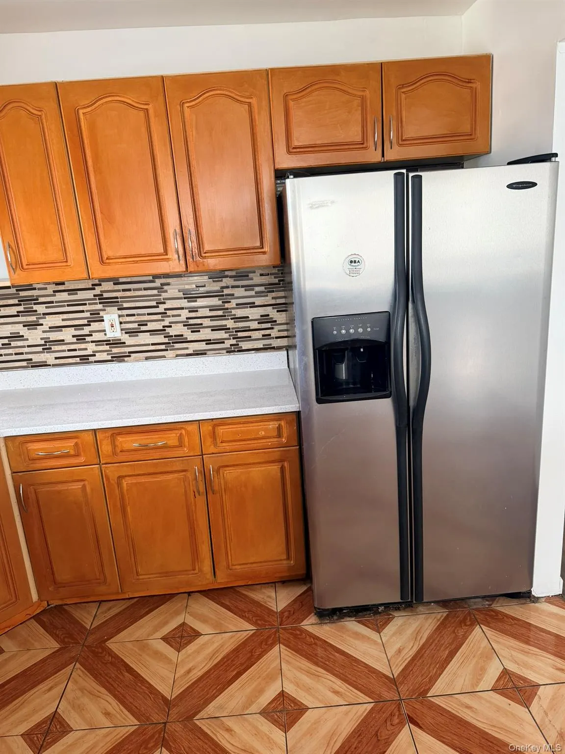 Wood-finish cabinetry, stainless steel side-by-side refrigerator, patterned tile flooring, and white countertops Wood-finish cabinetry, stainless steel side-by-side refrigerator, patterned tile flooring, and white countertops
