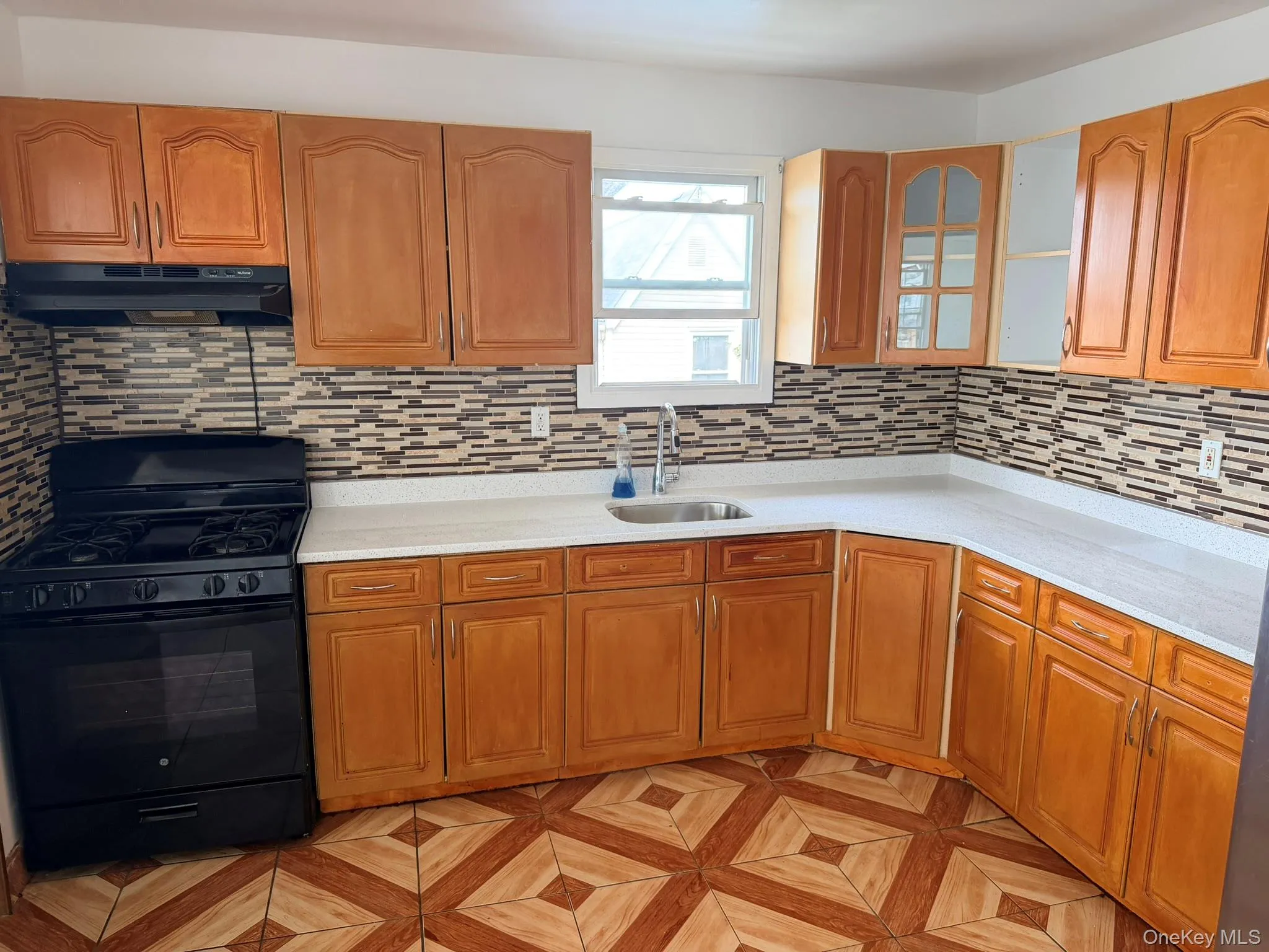 Kitchen featuring wood-finish cabinetry, light-toned countertops, a mosaic tile backsplash, and patterned floor tiling Kitchen featuring wood-finish cabinetry, light-toned countertops, a mosaic tile backsplash, and patterned floor tiling