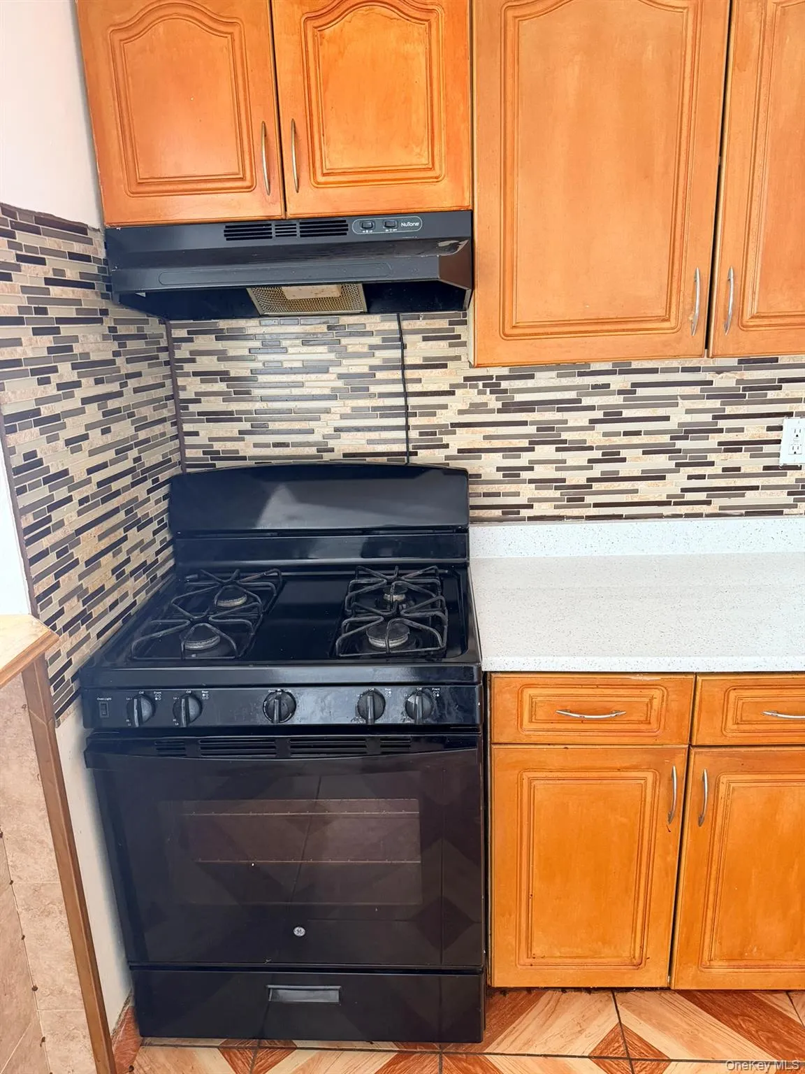 Black gas range with an overhead exhaust fan, mosaic tile backsplash, wood-finish cabinetry, and light-toned countertop Black gas range with an overhead exhaust fan, mosaic tile backsplash, wood-finish cabinetry, and light-toned countertop