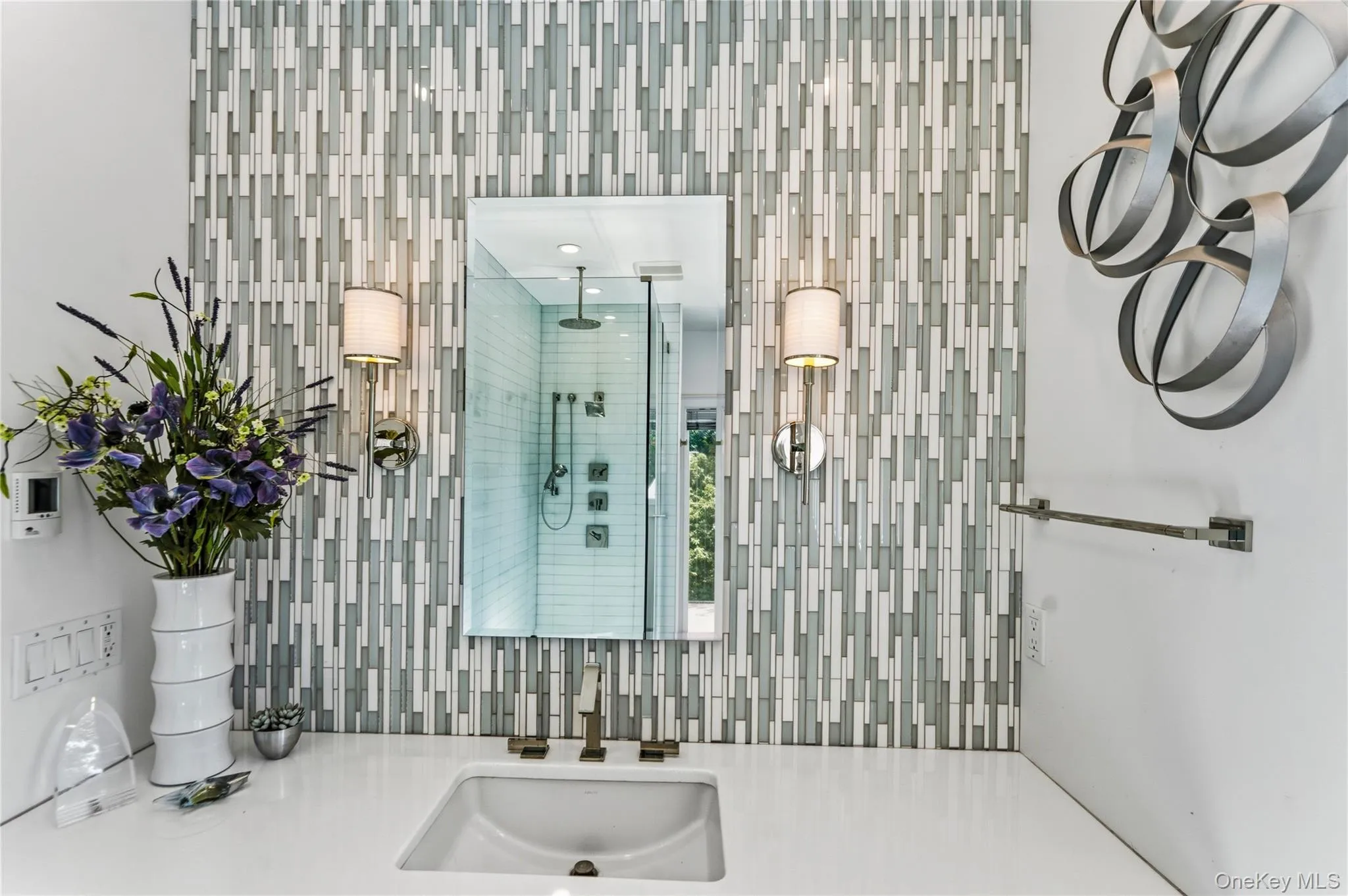 Vertical mosaic tile backsplash, polished countertop with an integrated rectangular sink, wall-mounted brushed metal faucet, dual sconce lighting, and a frameless mirror Vertical mosaic tile backsplash, polished countertop with an integrated rectangular sink, wall-mounted brushed metal faucet, dual sconce lighting, and a frameless mirror
