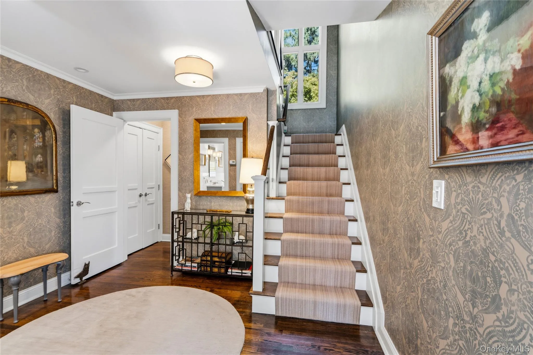 Foyer featuring patterned wallpaper, dark-stained wood flooring, a staircase with a central runner, a multi-pane fixed window, and crisp white millwork including paneled doors Foyer featuring patterned wallpaper, dark-stained wood flooring, a staircase with a central runner, a multi-pane fixed window, and crisp white millwork including paneled doors