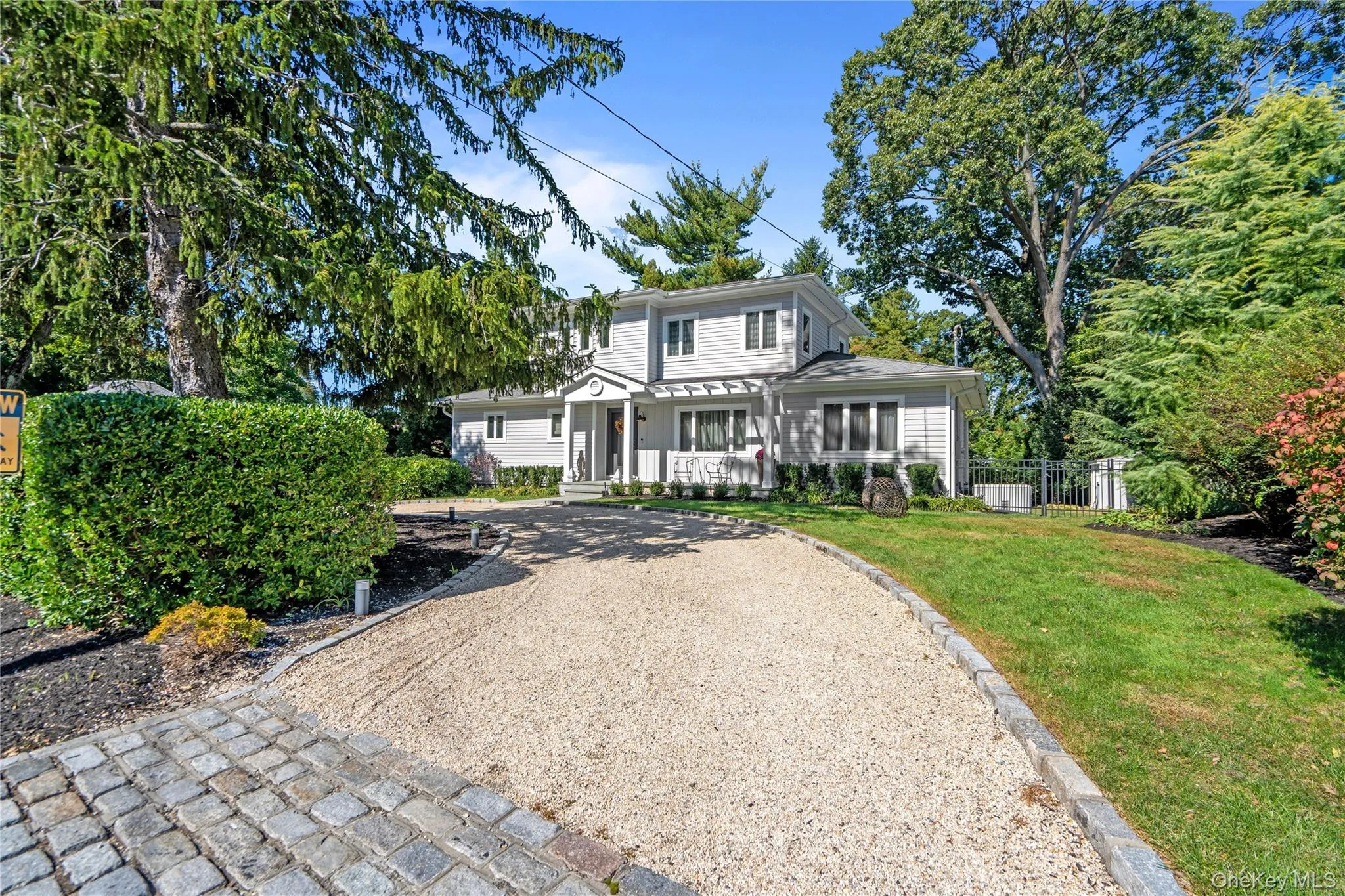 Two-story exterior featuring light-toned horizontal siding, a covered entryway with a pediment, a gravel driveway, cobble-style apron, and mature landscaping Two-story exterior featuring light-toned horizontal siding, a covered entryway with a pediment, a gravel driveway, cobble-style apron, and mature landscaping