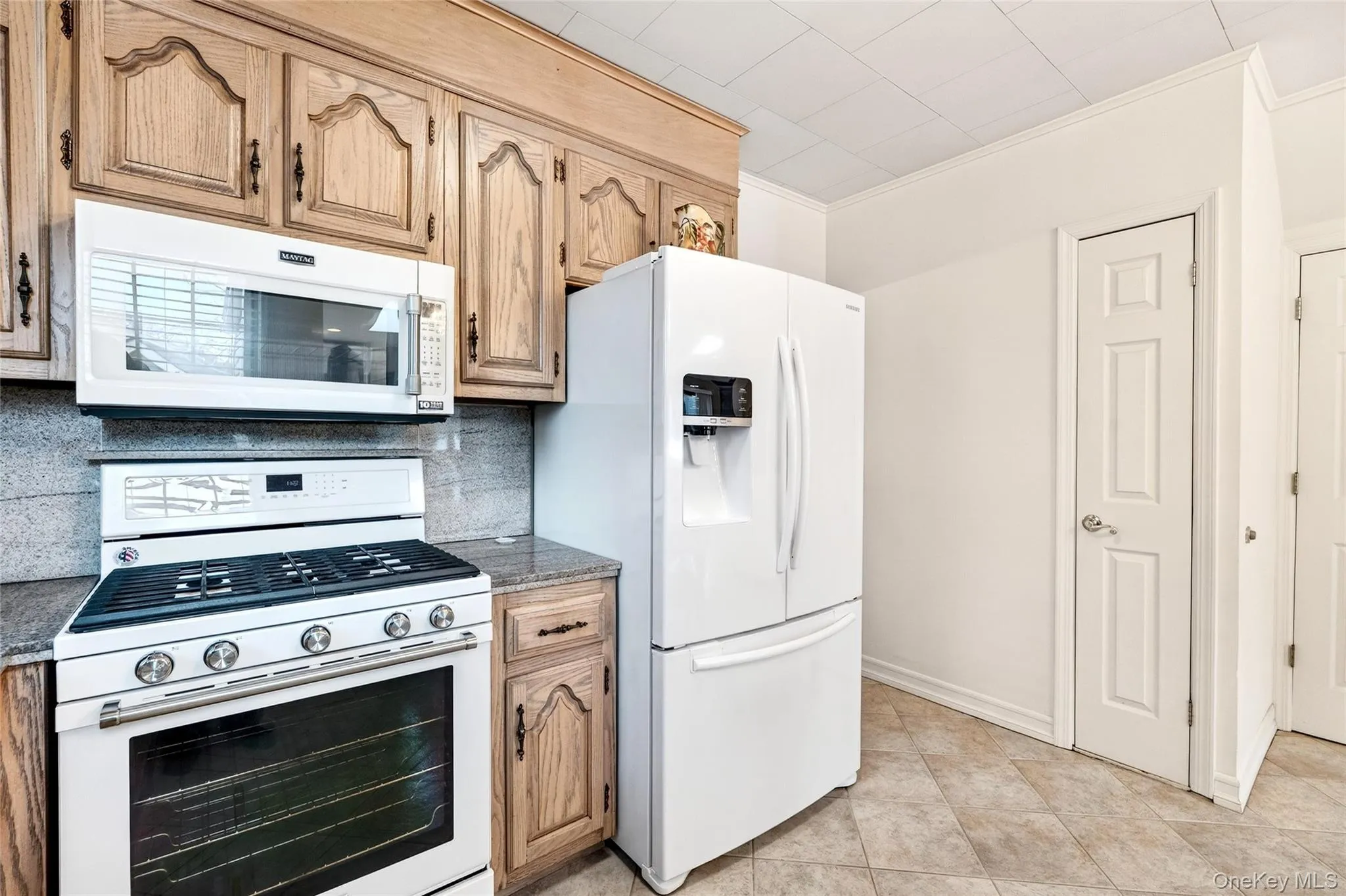 Kitchen featuring white appliances, ornamental molding, backsplash, dark stone countertops, and light brown cabinetry Kitchen featuring white appliances, ornamental molding, backsplash, dark stone countertops, and light brown cabinetry