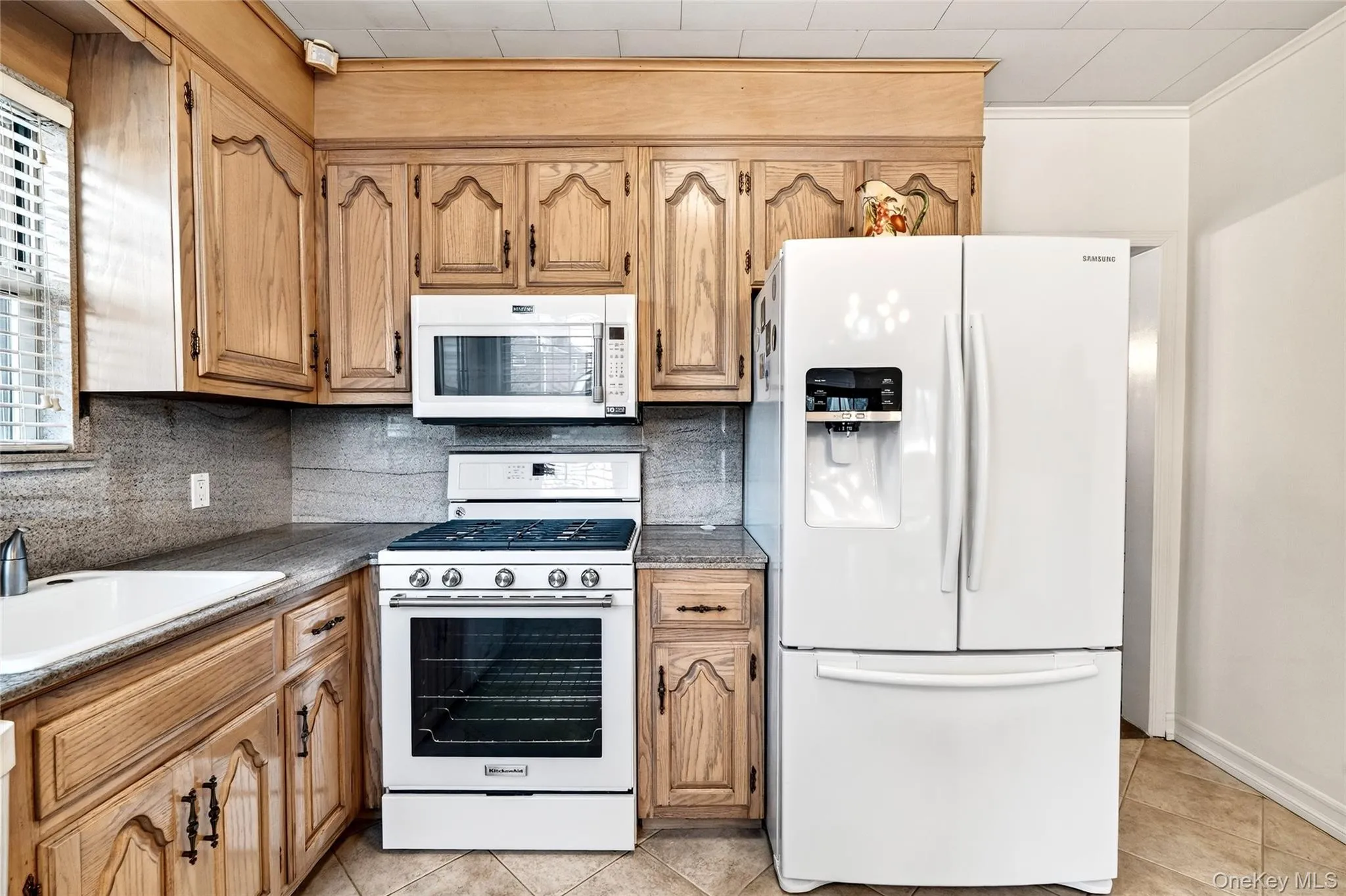Kitchen with white appliances, backsplash, light tile patterned flooring, dark stone counters, and brown cabinets Kitchen with white appliances, backsplash, light tile patterned flooring, dark stone counters, and brown cabinets