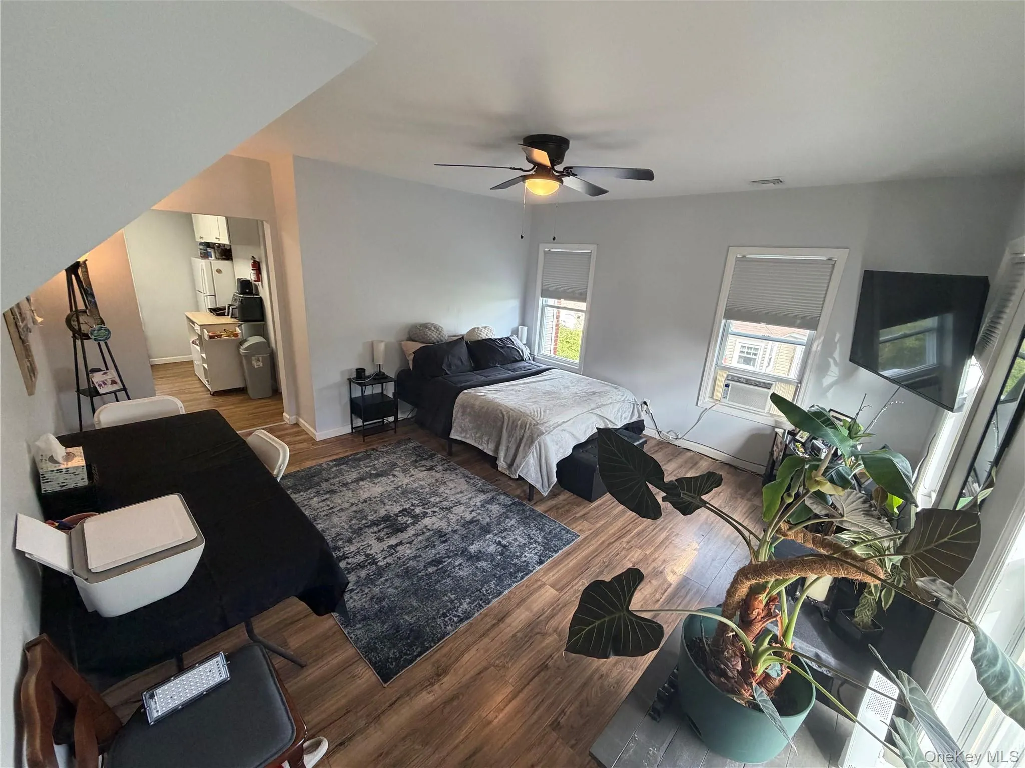 Wood-finish flooring throughout, ceiling fan, multiple windows with blinds, and a partial view of an adjacent kitchen space Wood-finish flooring throughout, ceiling fan, multiple windows with blinds, and a partial view of an adjacent kitchen space