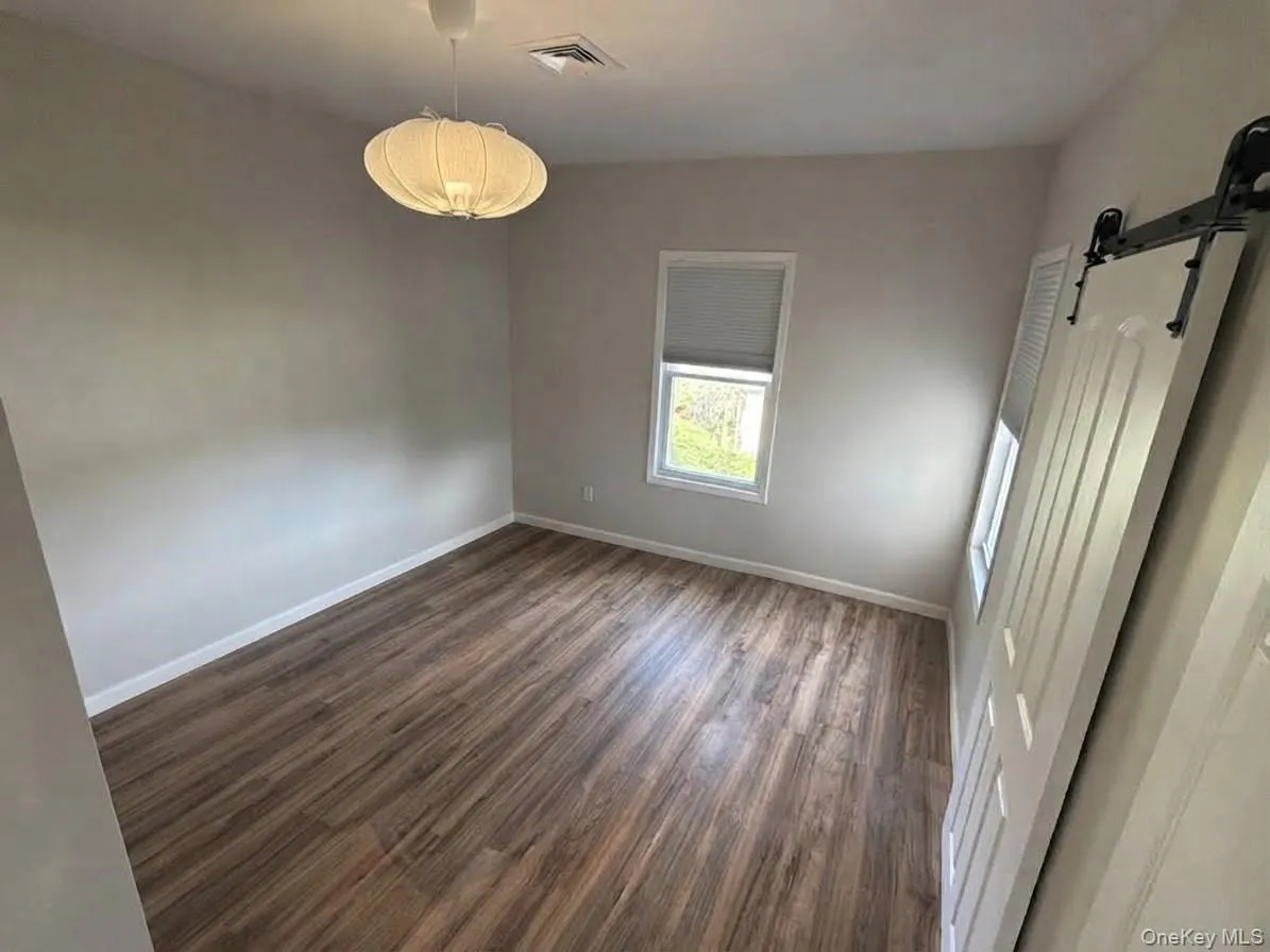Wood-finish flooring, single window with blinds, overhead lighting, and a white barn door Wood-finish flooring, single window with blinds, overhead lighting, and a white barn door