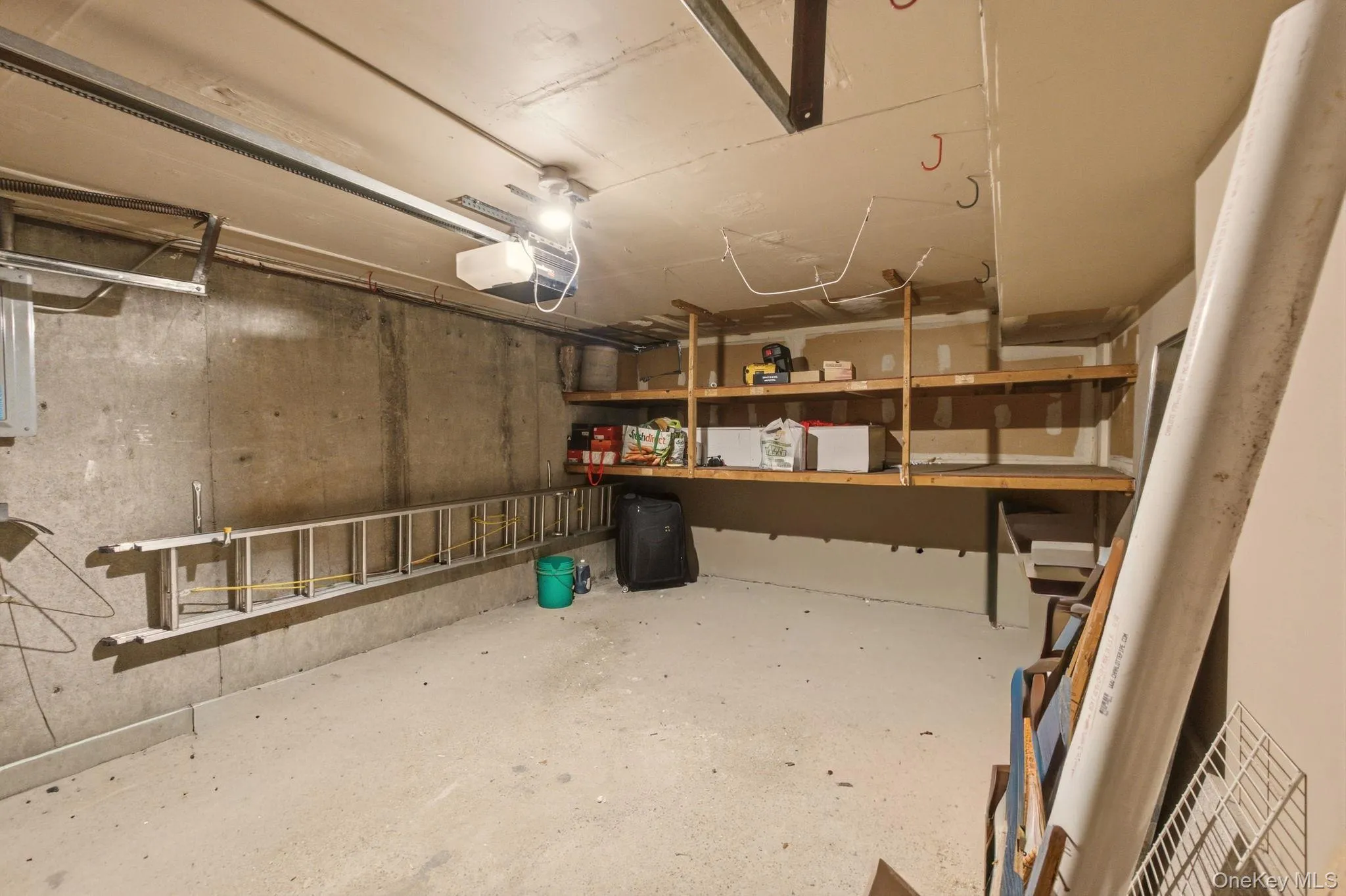 Garage space featuring concrete walls, built-in wooden shelving, and an overhead garage door mechanism Garage space featuring concrete walls, built-in wooden shelving, and an overhead garage door mechanism
