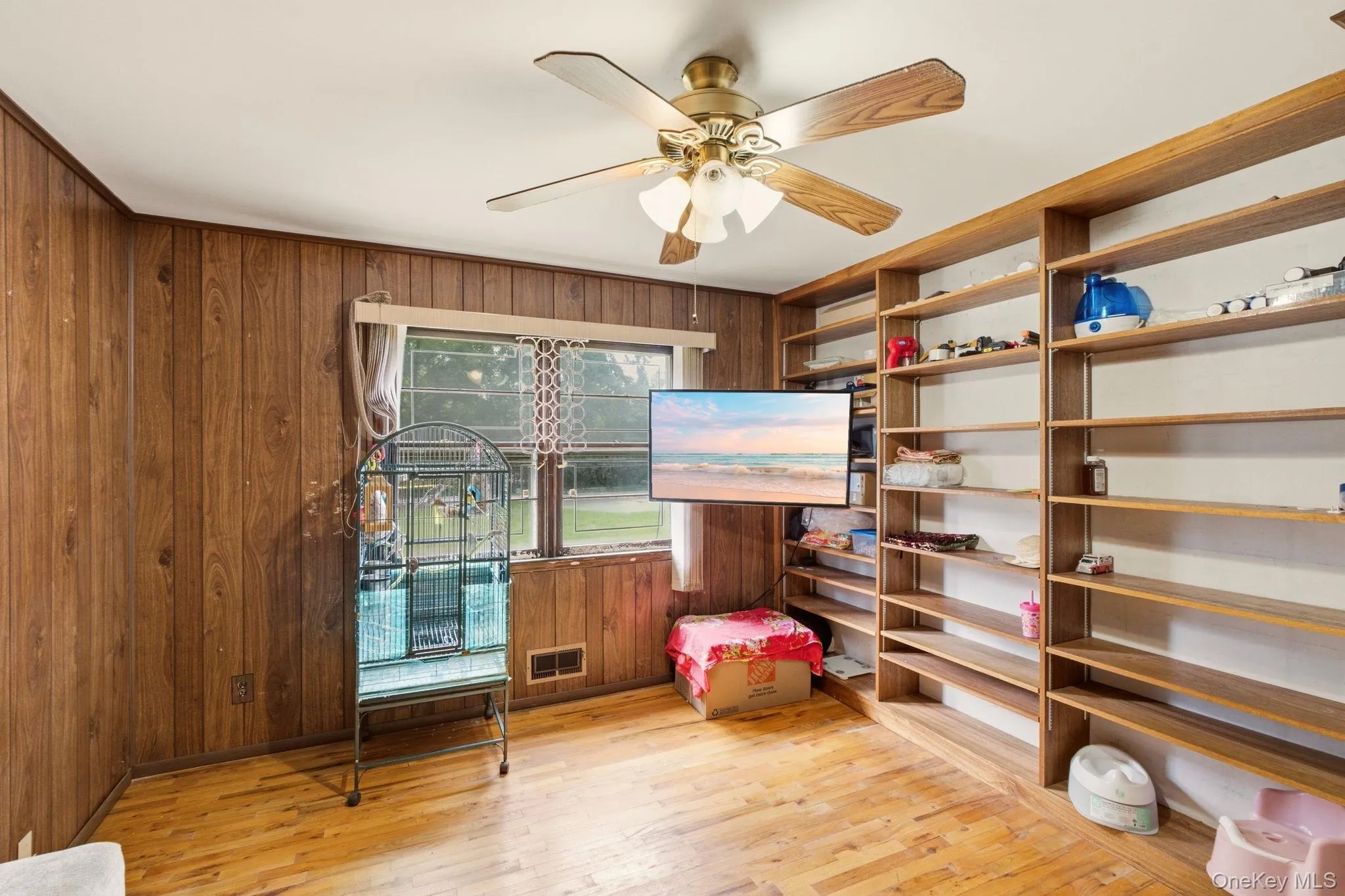 Wood-finish flooring, wood paneling, built-in shelving, ceiling fan with light fixture, and window with natural light Wood-finish flooring, wood paneling, built-in shelving, ceiling fan with light fixture, and window with natural light