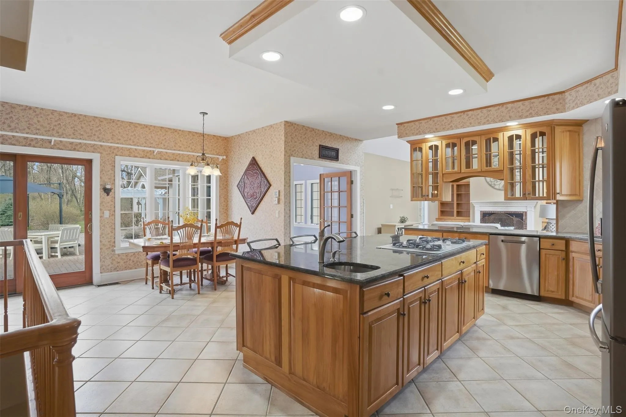The kitchen features light wood cabinetry, a central island with a dark countertop and integrated sink, stainless steel appliances, and tiled flooring The kitchen features light wood cabinetry, a central island with a dark countertop and integrated sink, stainless steel appliances, and tiled flooring