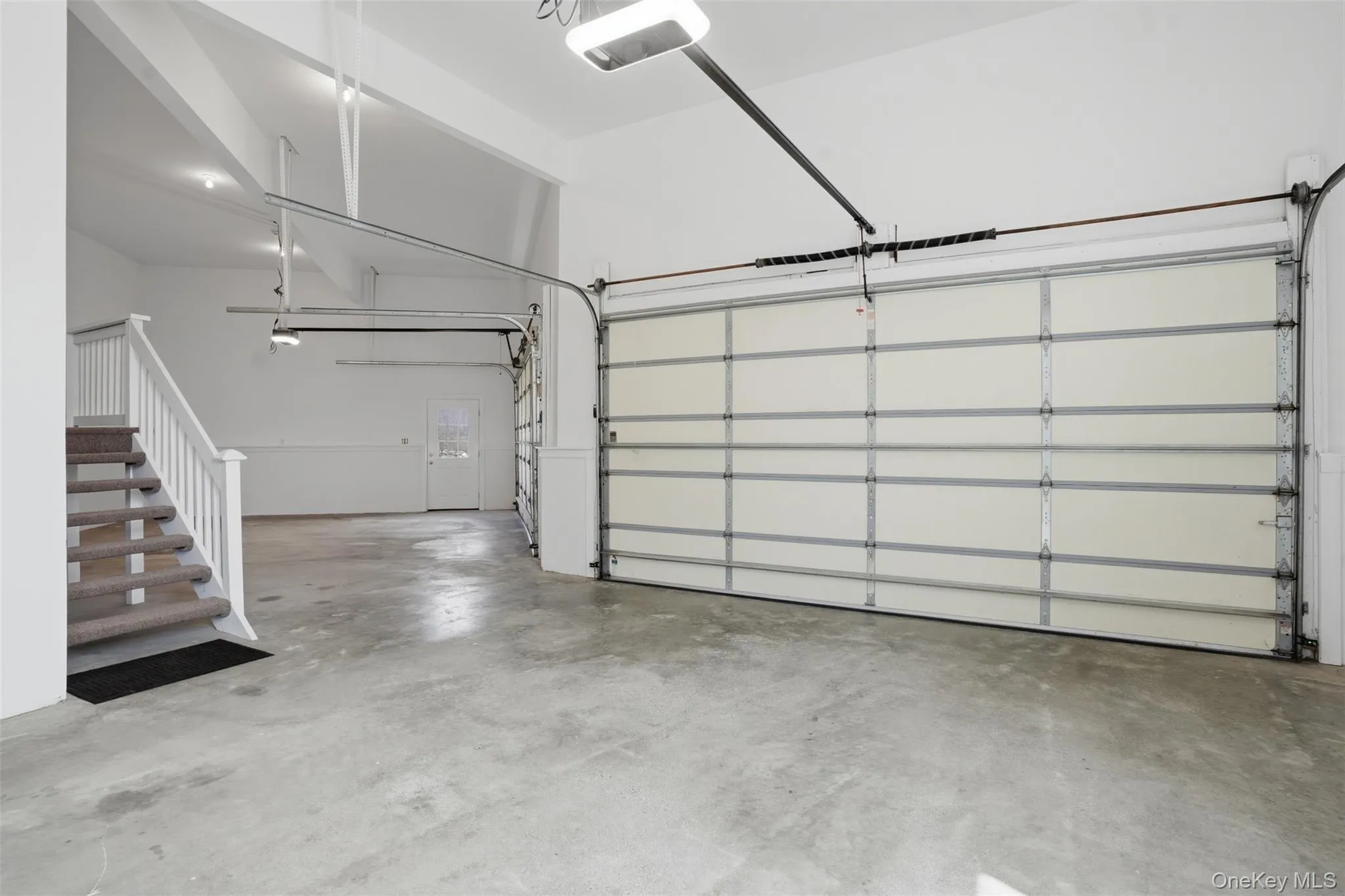 The property features an expansive garage with concrete flooring, a white automatic garage door, and a staircase with carpeted treads and white railings The property features an expansive garage with concrete flooring, a white automatic garage door, and a staircase with carpeted treads and white railings