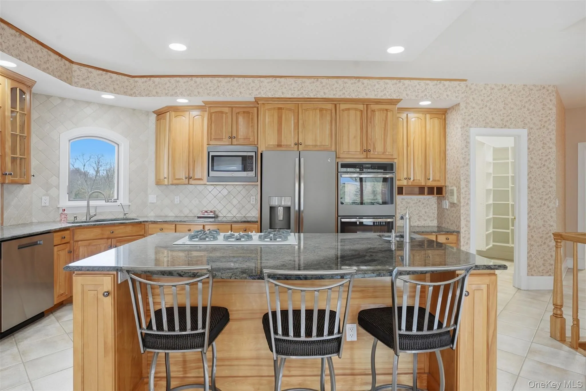 The kitchen features wood cabinetry, stainless steel appliances, a large island with a cooktop and seating, granite countertops, and a tiled backsplash The kitchen features wood cabinetry, stainless steel appliances, a large island with a cooktop and seating, granite countertops, and a tiled backsplash