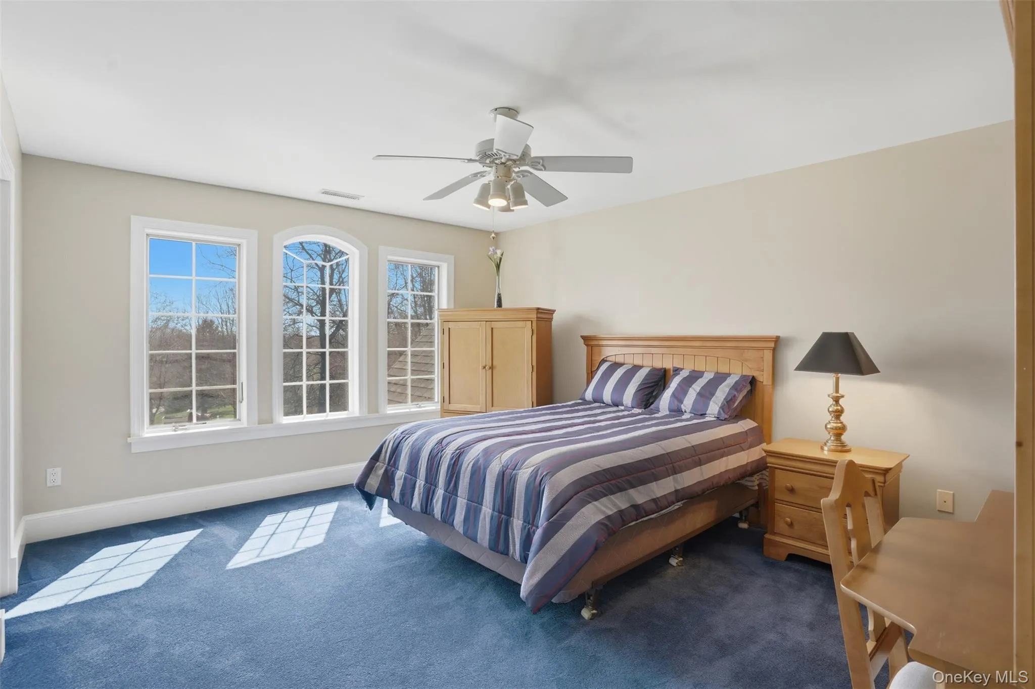 This bright bedroom features a ceiling fan, plush blue carpet, and a trio of windows offering views of the outdoors This bright bedroom features a ceiling fan, plush blue carpet, and a trio of windows offering views of the outdoors