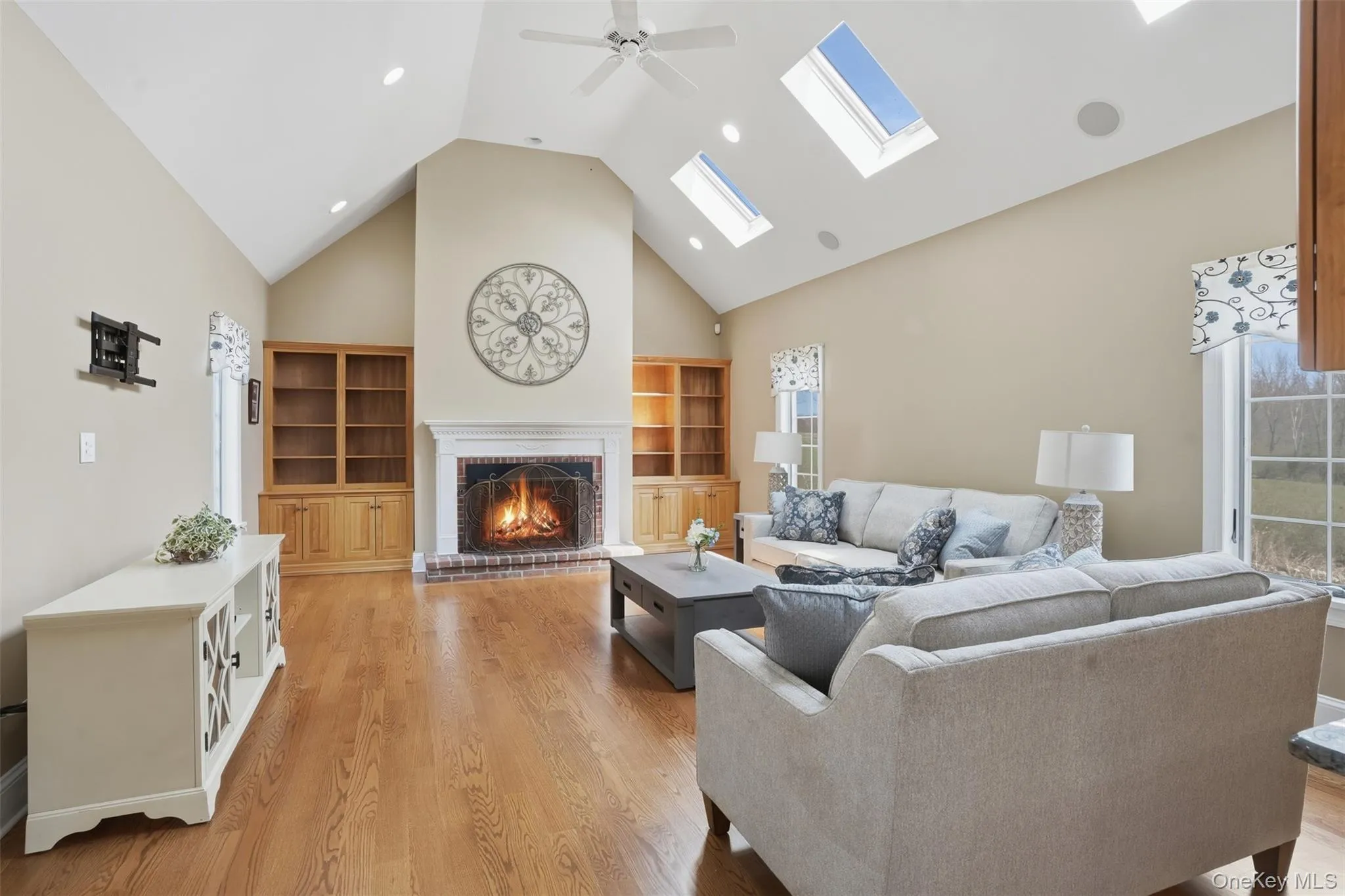Spacious living area featuring vaulted ceilings, hardwood floors, a brick fireplace with a white mantel, built-in wooden shelving, and skylights providing abundant natural light Spacious living area featuring vaulted ceilings, hardwood floors, a brick fireplace with a white mantel, built-in wooden shelving, and skylights providing abundant natural light
