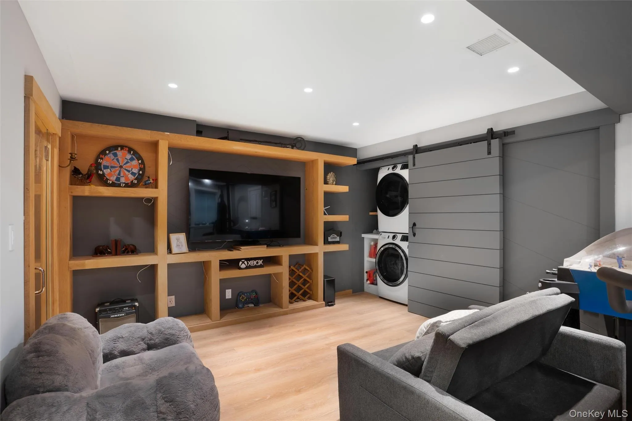 Built-in entertainment center with open shelving, light wood-finish flooring, recessed lighting, and a barn door concealing stacked laundry appliances Built-in entertainment center with open shelving, light wood-finish flooring, recessed lighting, and a barn door concealing stacked laundry appliances