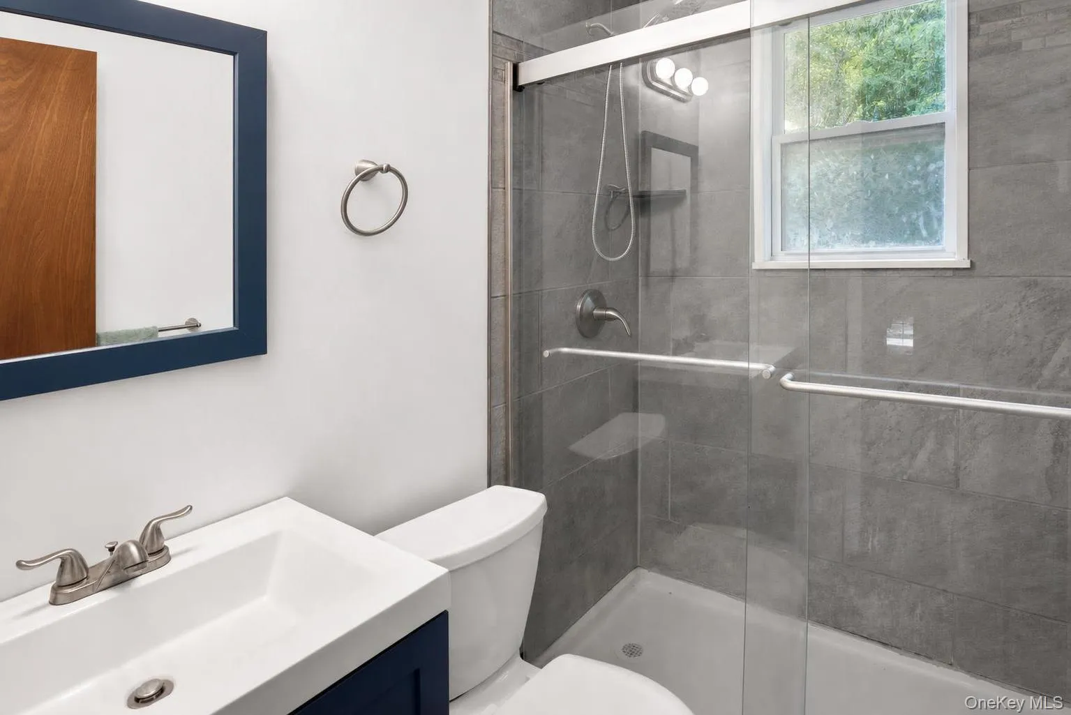 Bathroom featuring a vanity with an integrated sink, a toilet, a shower with a sliding glass door, grey tile surround, and a window Bathroom featuring a vanity with an integrated sink, a toilet, a shower with a sliding glass door, grey tile surround, and a window