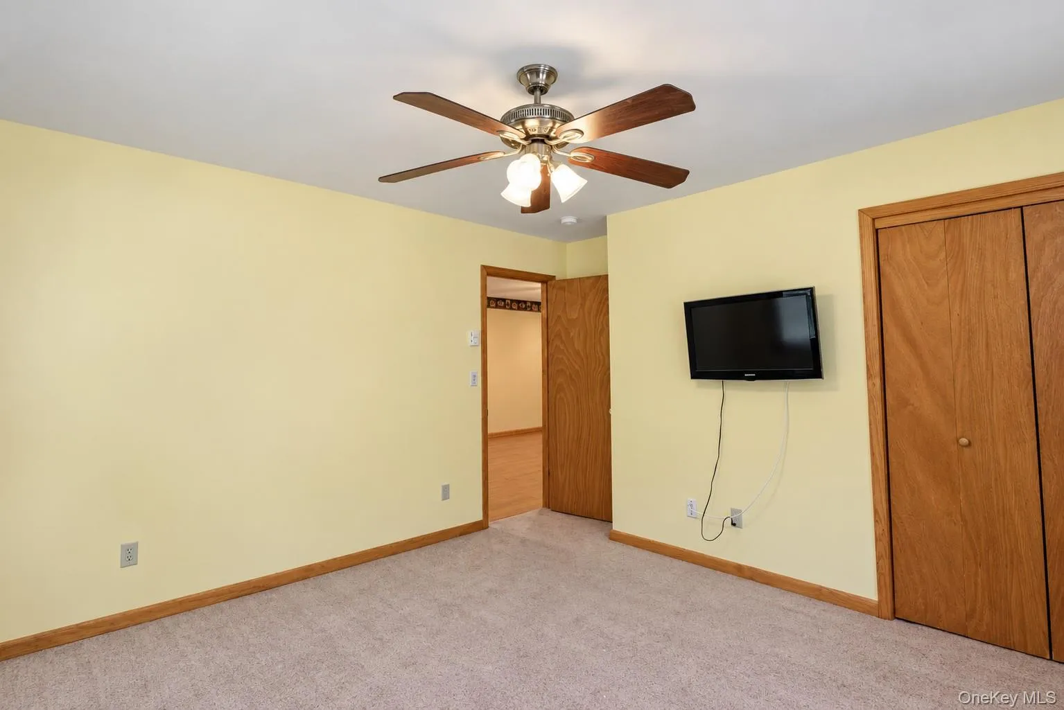 Ceiling fan with integrated lighting, wood-finish bi-fold closet doors, wall-mounted television, and neutral carpeting Ceiling fan with integrated lighting, wood-finish bi-fold closet doors, wall-mounted television, and neutral carpeting