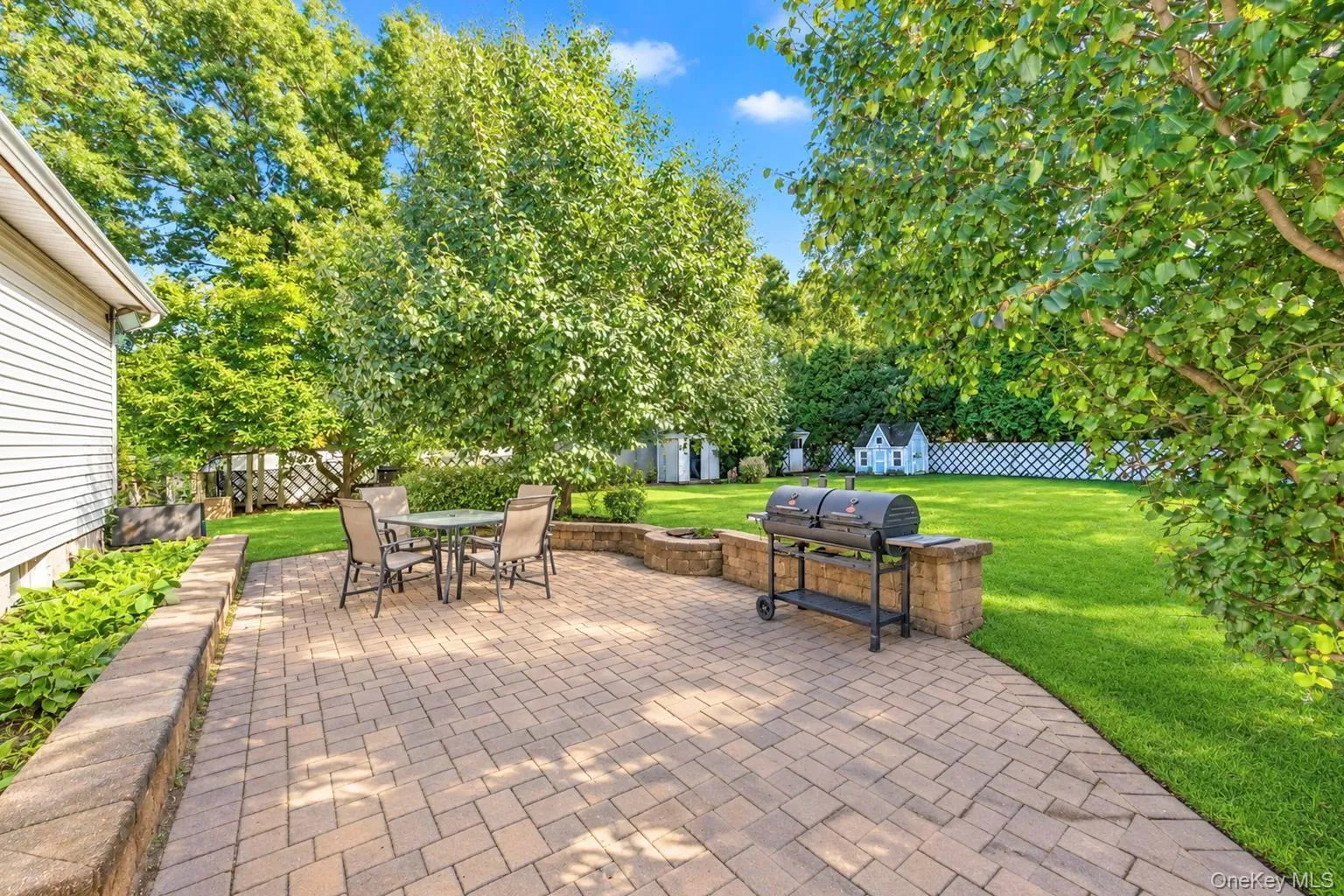 Expansive backyard featuring a paver patio with a built-in stone-look planter, a separate stone-look grilling station, and a manicured lawn surrounded by mature trees Expansive backyard featuring a paver patio with a built-in stone-look planter, a separate stone-look grilling station, and a manicured lawn surrounded by mature trees