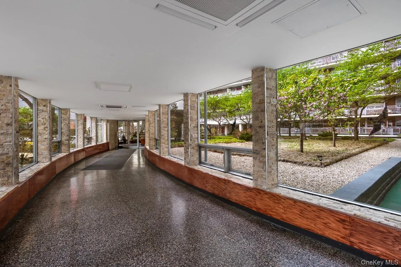 Curved corridor featuring terrazzo flooring, stone-clad columns, and floor-to-ceiling windows overlooking a landscaped courtyard with trees and a water feature Curved corridor featuring terrazzo flooring, stone-clad columns, and floor-to-ceiling windows overlooking a landscaped courtyard with trees and a water feature