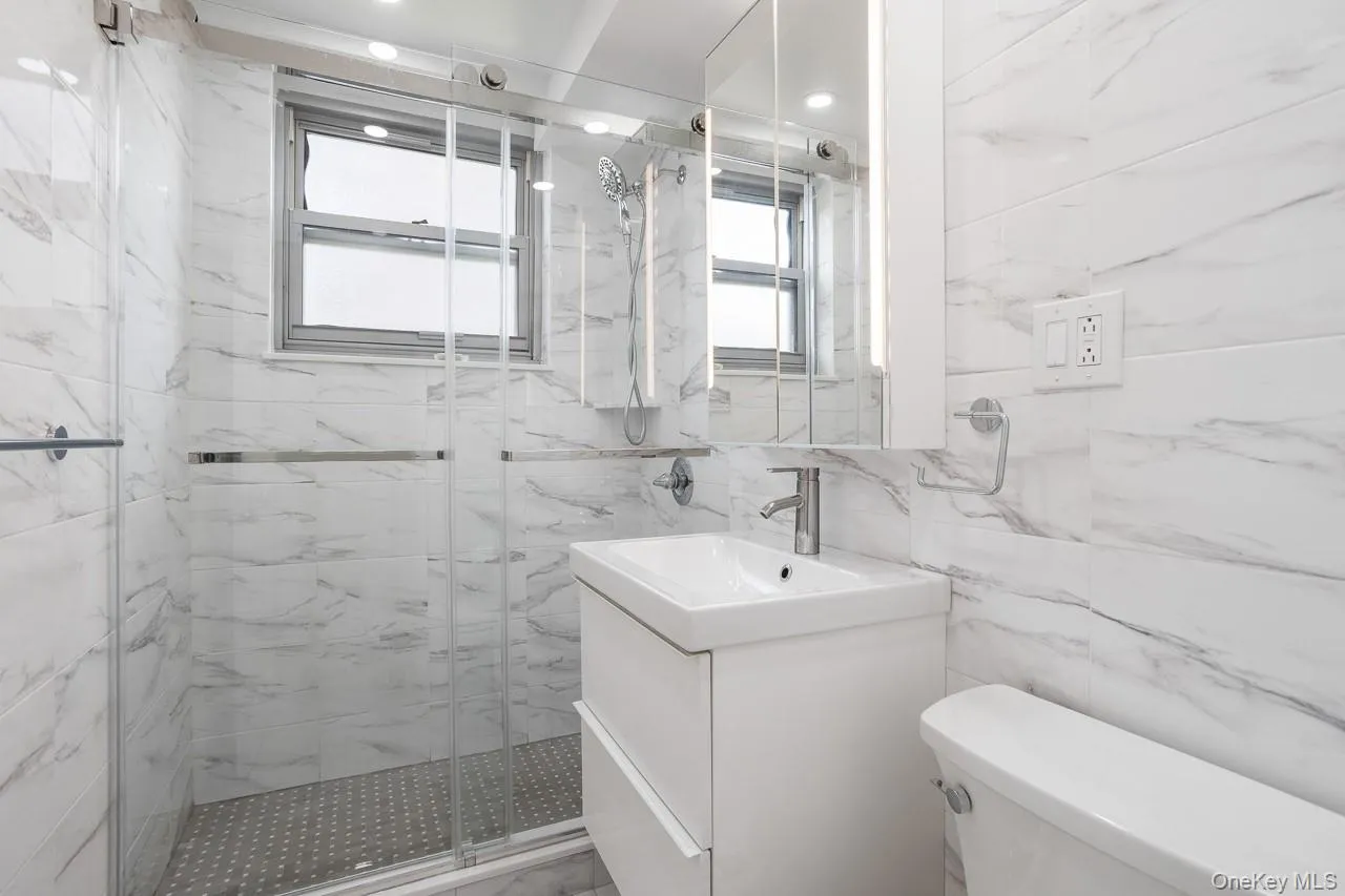 Walk-in shower with clear glass enclosure, white vanity with integrated sink, wall-mounted mirrored medicine cabinet with integrated lighting, window, and fully tiled walls Walk-in shower with clear glass enclosure, white vanity with integrated sink, wall-mounted mirrored medicine cabinet with integrated lighting, window, and fully tiled walls