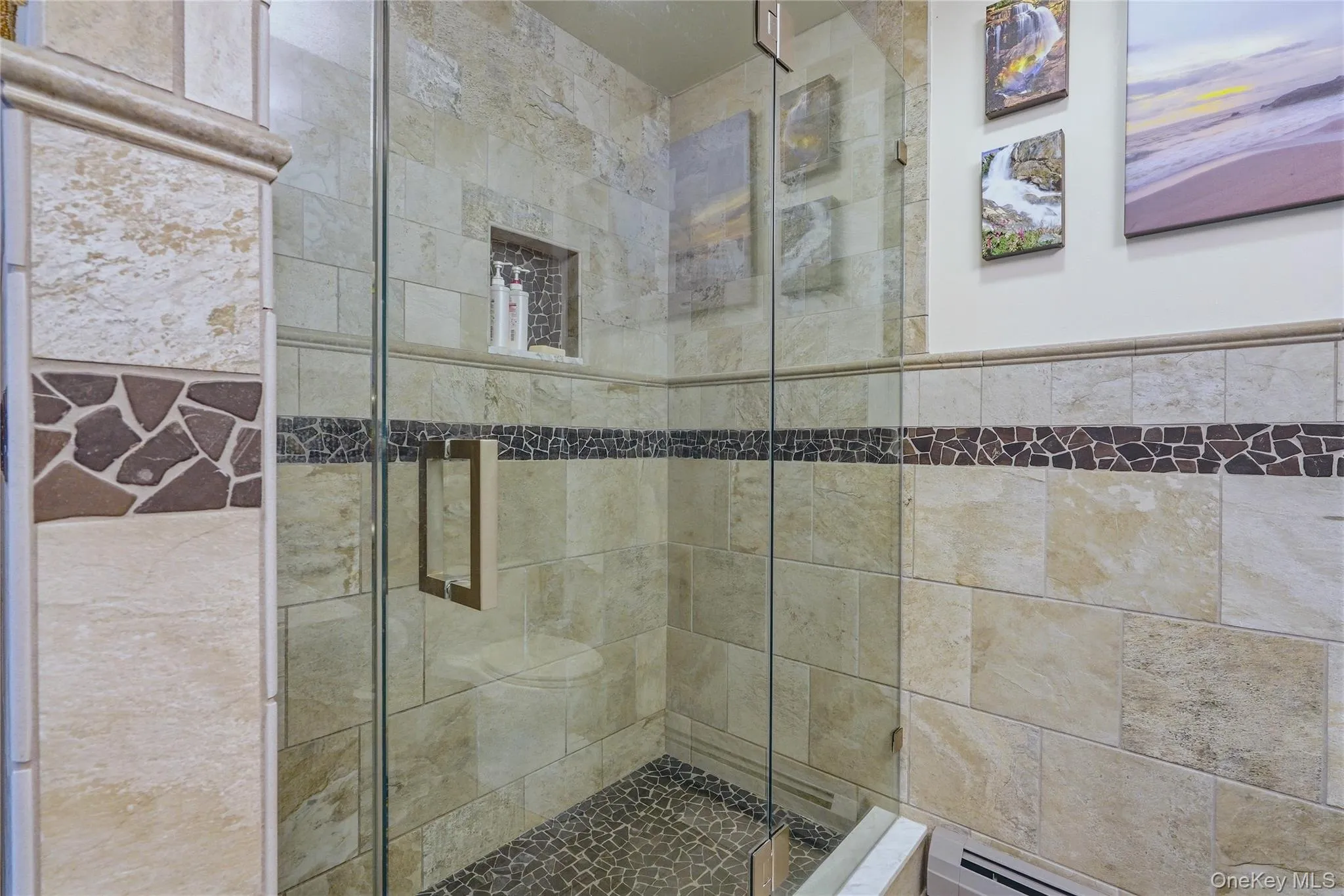 Glass-enclosed shower featuring natural stone-finish wall tile, a decorative mosaic tile band, a recessed shower niche, and a river rock-finish shower floor Glass-enclosed shower featuring natural stone-finish wall tile, a decorative mosaic tile band, a recessed shower niche, and a river rock-finish shower floor