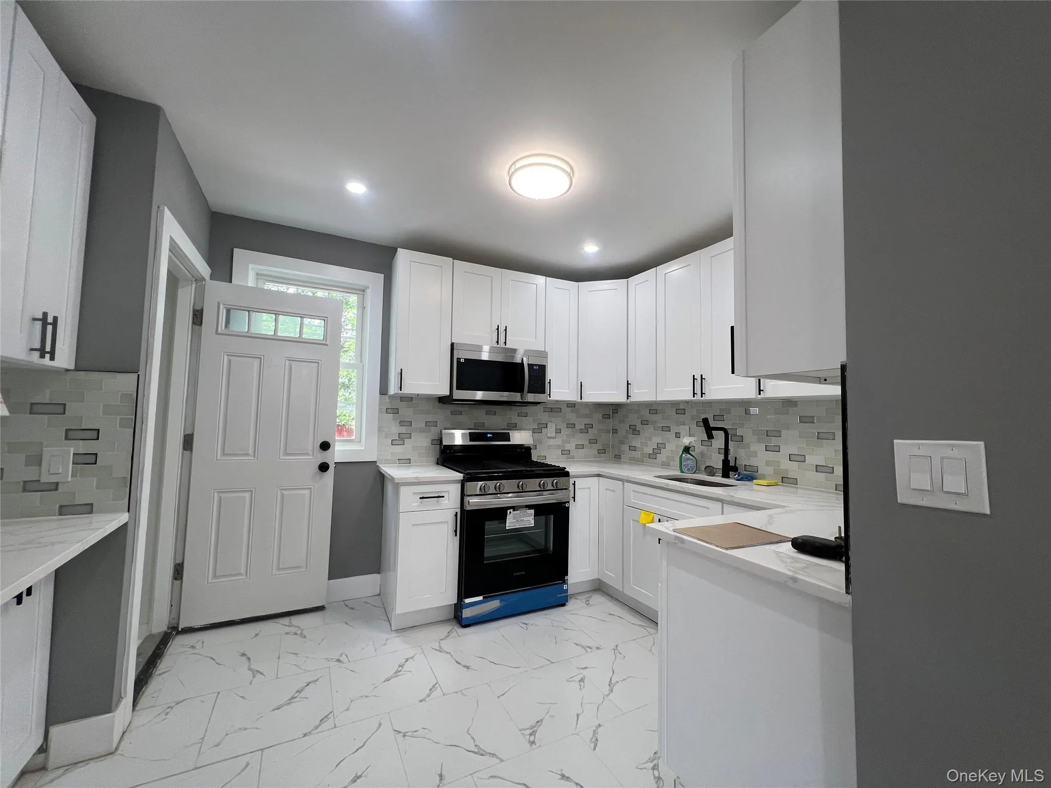 Modern kitchen featuring white shaker cabinetry, stainless steel appliances, a mosaic tile backsplash, light-toned countertops, and white tile flooring Modern kitchen featuring white shaker cabinetry, stainless steel appliances, a mosaic tile backsplash, light-toned countertops, and white tile flooring
