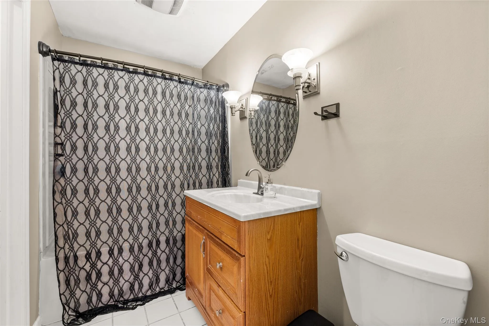 Oak finish vanity with a white countertop, oval mirror, wall-mounted light fixture, and white floor tiling Oak finish vanity with a white countertop, oval mirror, wall-mounted light fixture, and white floor tiling