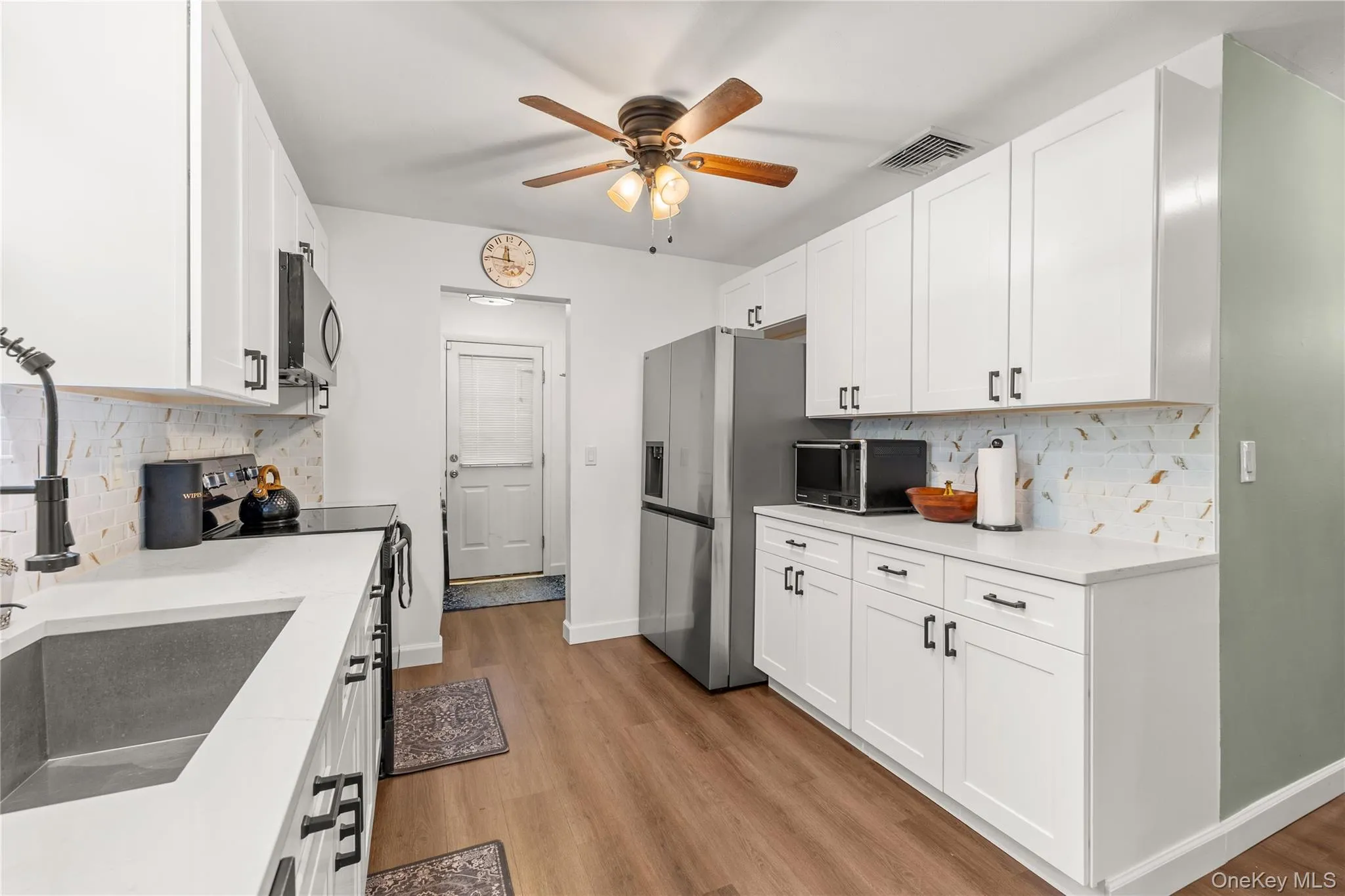 Kitchen featuring white shaker-style cabinetry, stainless steel appliances, wood-finish flooring, and light-toned countertops Kitchen featuring white shaker-style cabinetry, stainless steel appliances, wood-finish flooring, and light-toned countertops