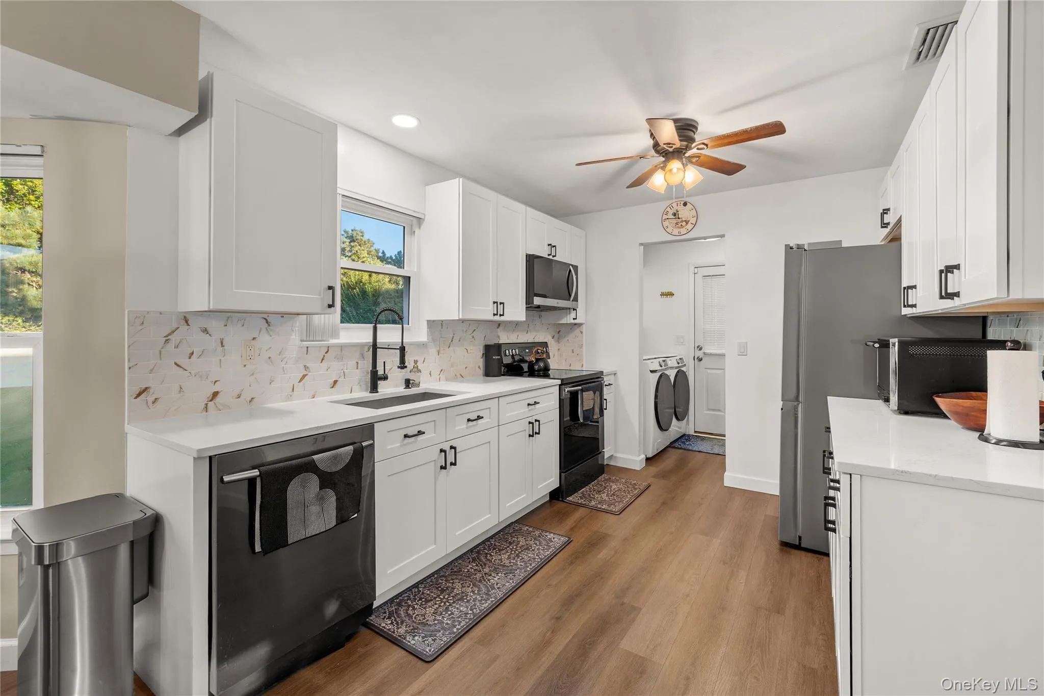 Kitchen featuring white shaker cabinetry, white quartz countertops, a decorative tile backsplash, stainless steel appliances, and wood-finish flooring. Laundry room attached to kitchen. Kitchen featuring white shaker cabinetry, white quartz countertops, a decorative tile backsplash, stainless steel appliances, and wood-finish flooring. Laundry room attached to kitchen.